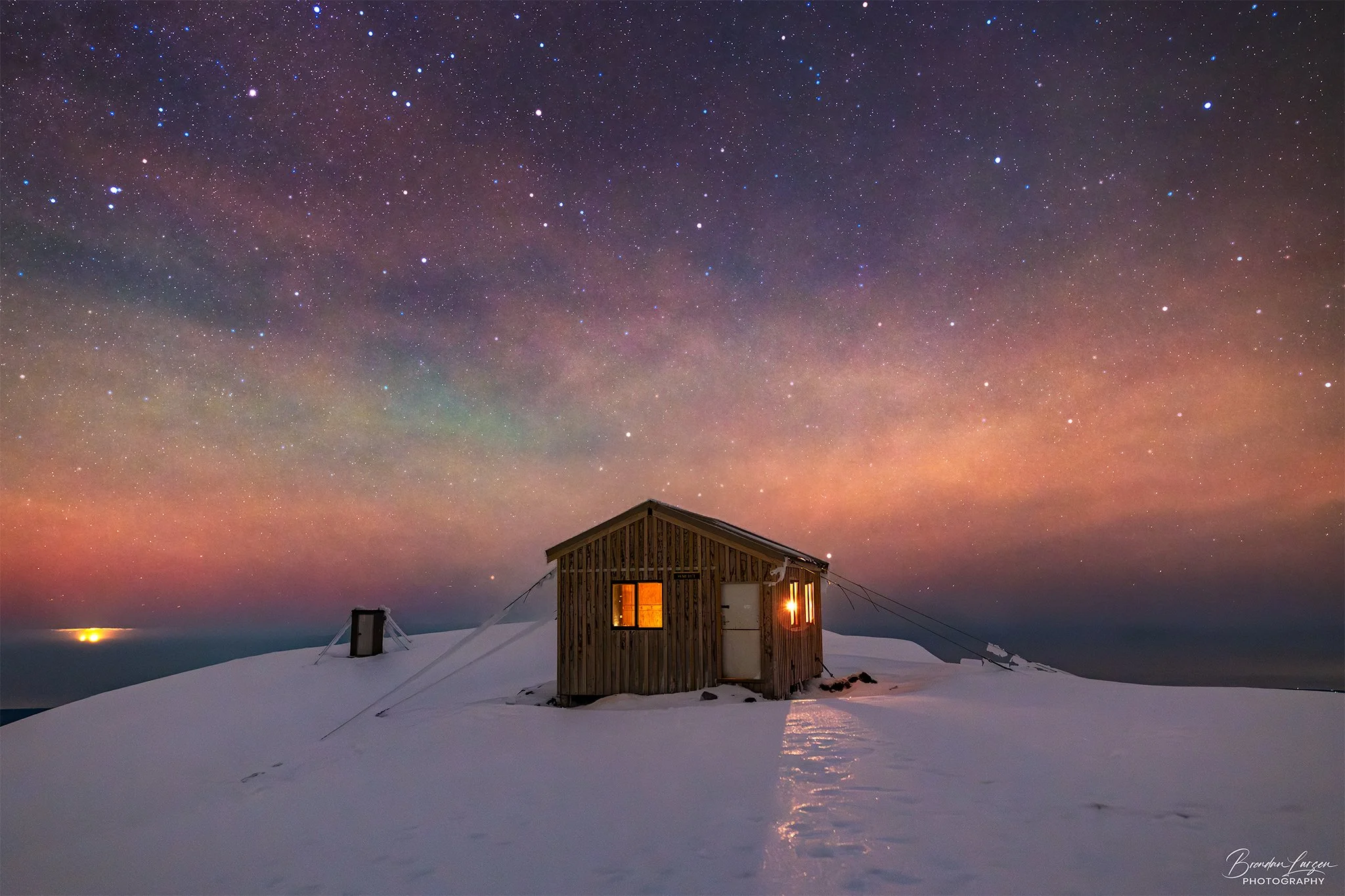 A wooden cabin with warm lights on a snowy landscape under a colorful starry night sky with visible the Milky Way galaxy.