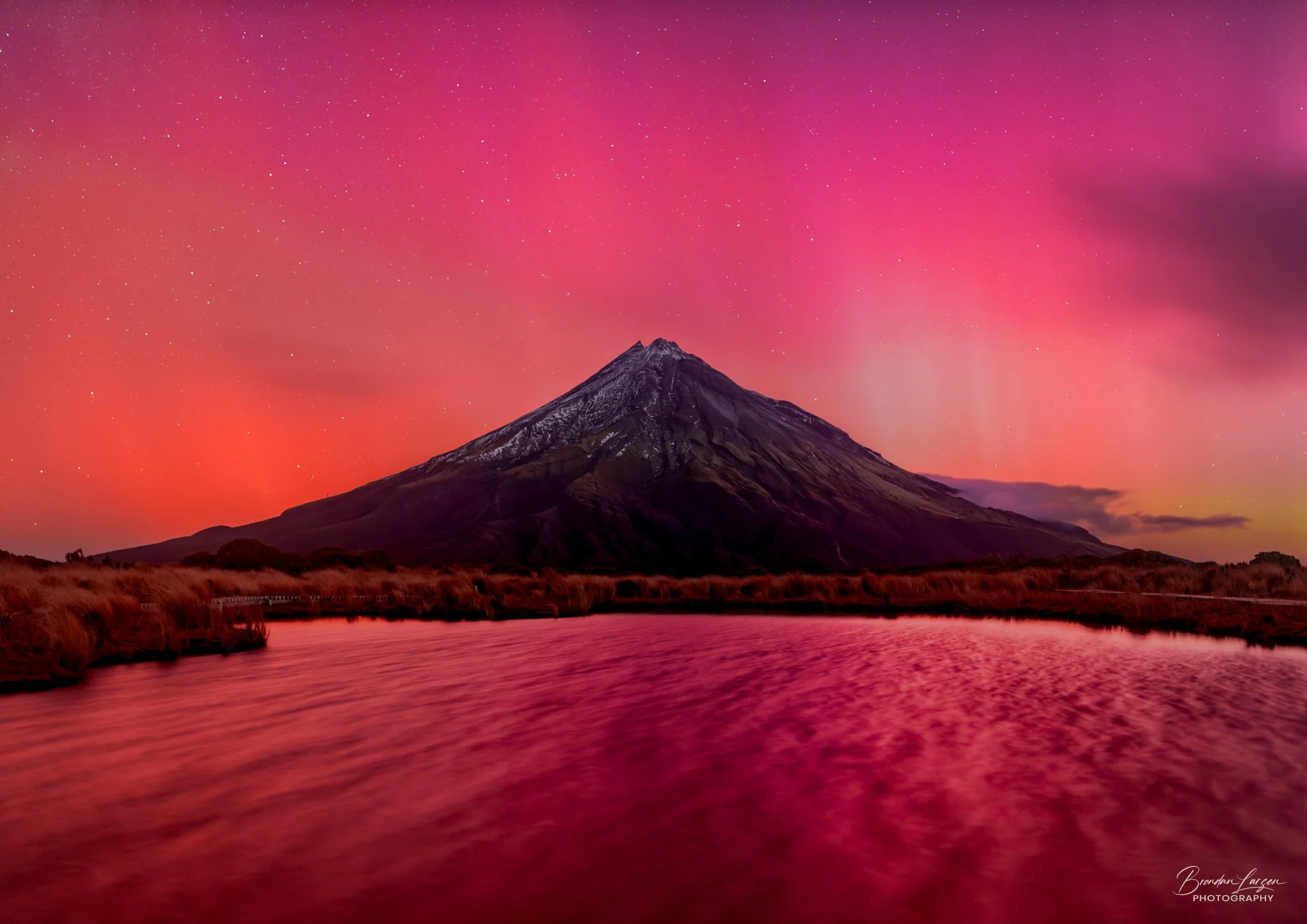 Snow-capped mountain under an aurora filled colorful, pink and purple starry sky, with a lake in the foreground reflecting the sky's colors.