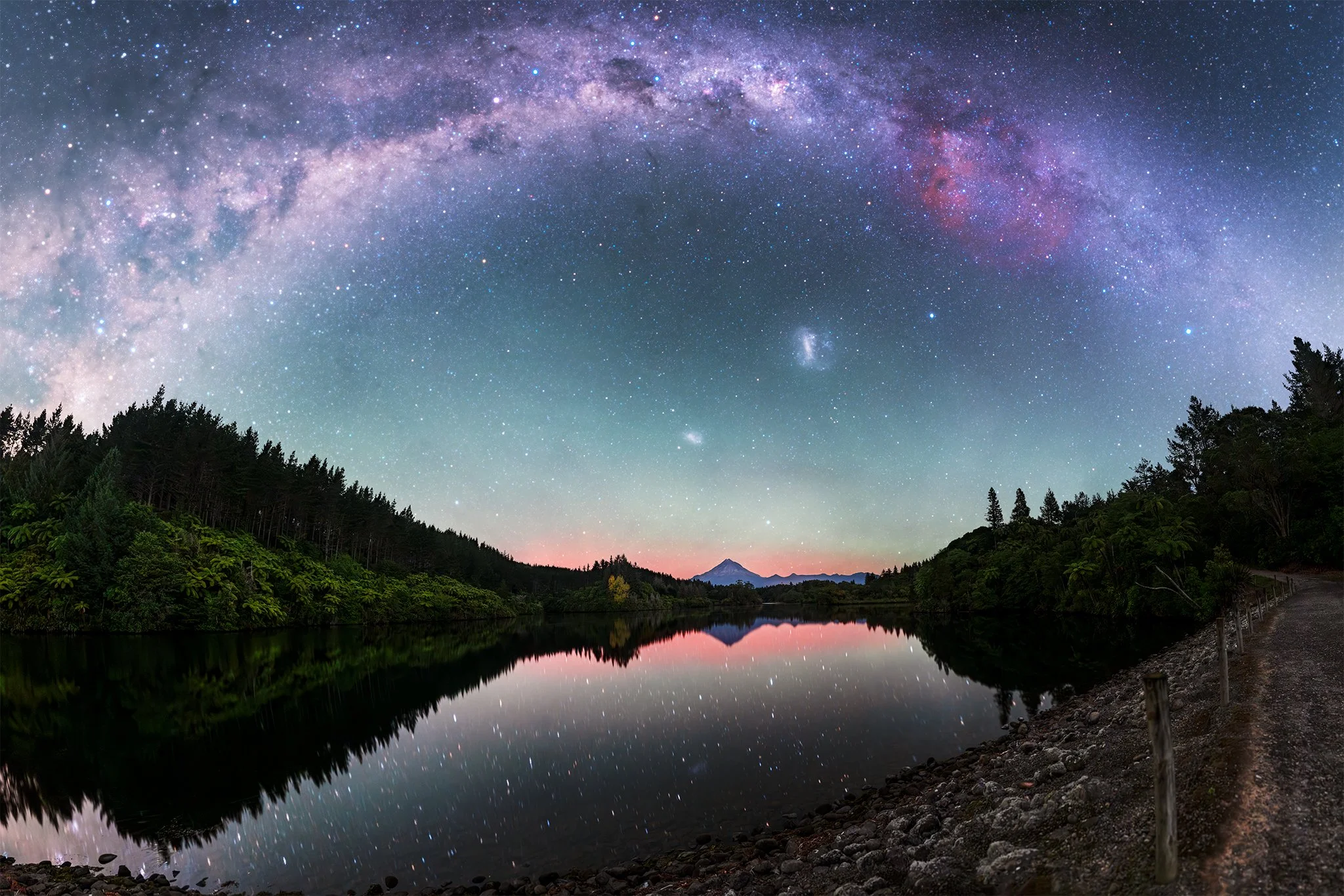Night sky with the Milky Way galaxy arcing over a calm lake, surrounded by green trees and a mountain, with a faint pink aurora and blue glow near the horizon.