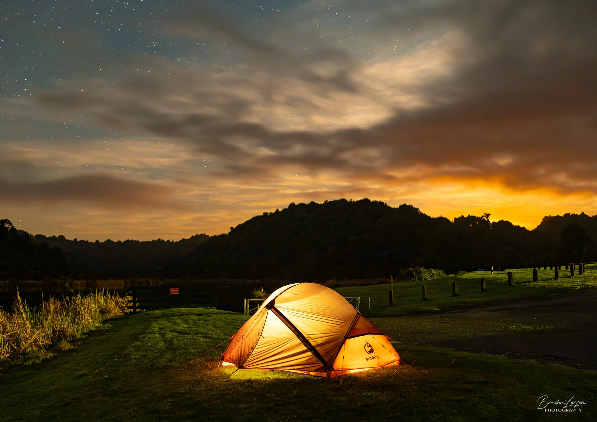 A glowing orange camping tent set up on grass near a lake at dusk, with a dark hillside in the background under a cloudy sky with stars visible.