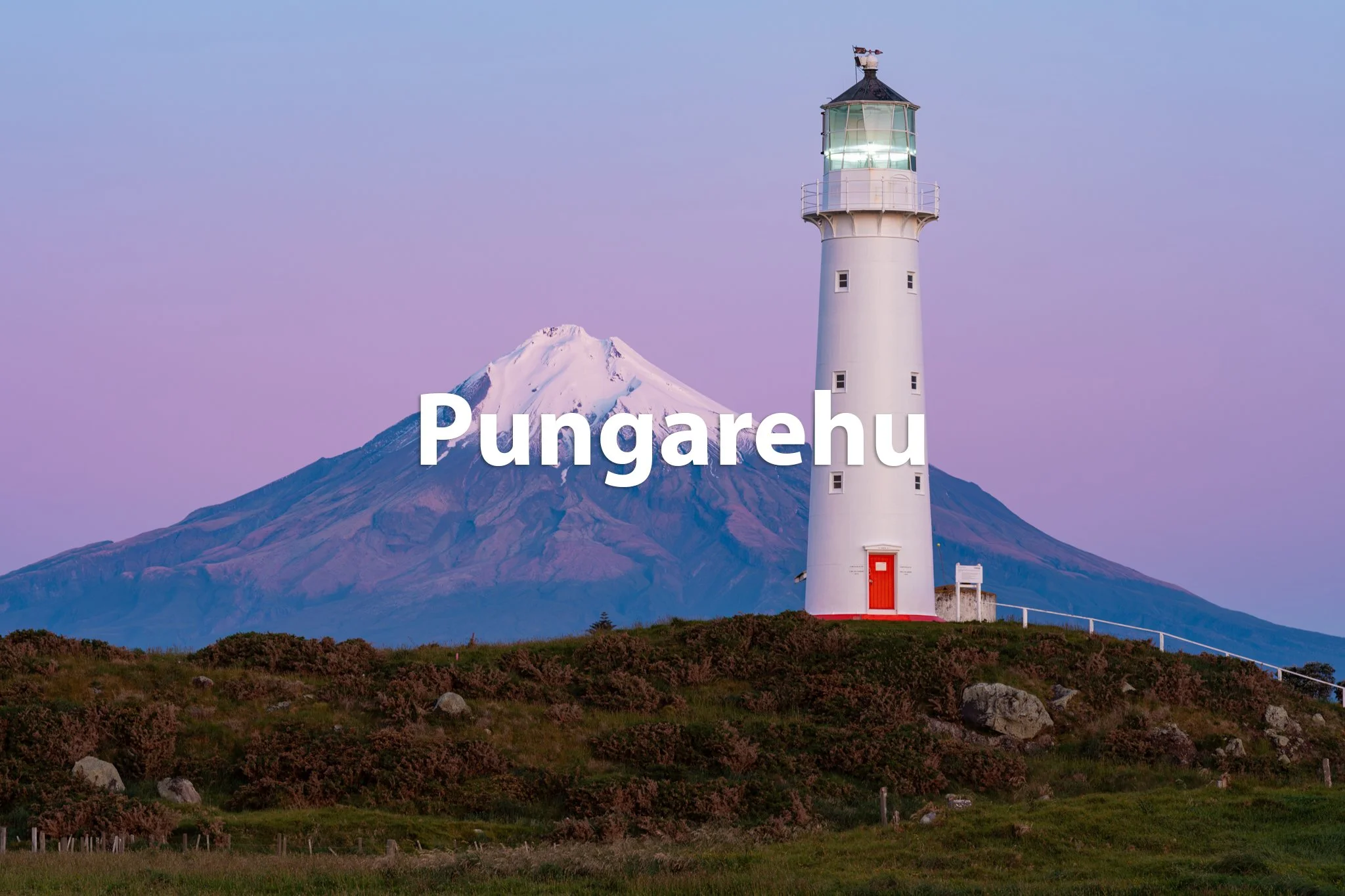 A white lighthouse with a red door and railing, positioned on a grassy hill, with a snow-capped mountain in the background under a pastel-colored sky.