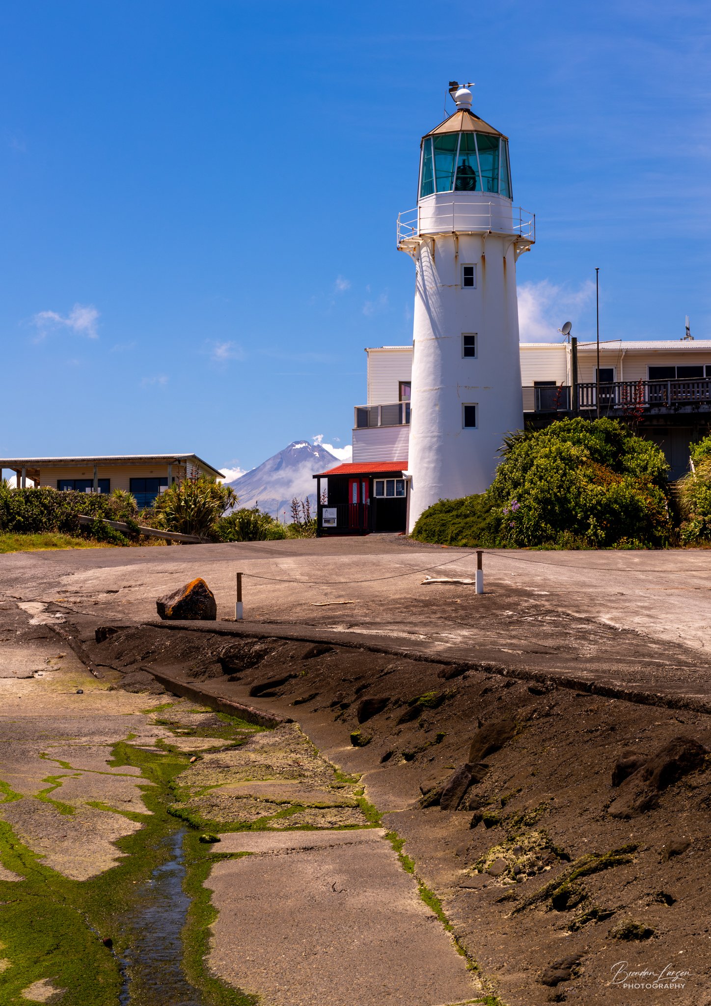 Lighthouse near the coast with Mount Taranaki volcano in the background under a blue sky.