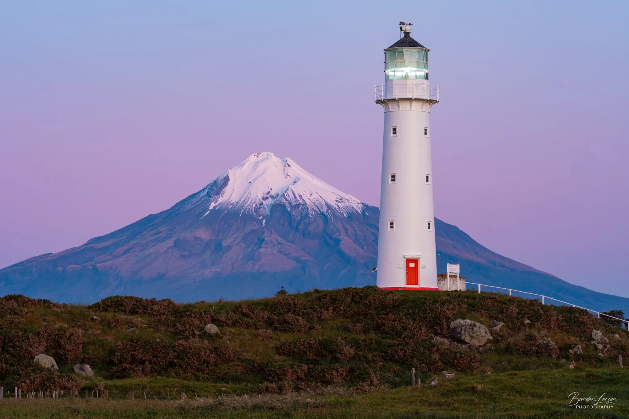 A tall white lighthouse with a red door standing on a grassy hill, with Mount Taranaki (Mount Egmont) snow-capped volcano in the background under a pastel sky.