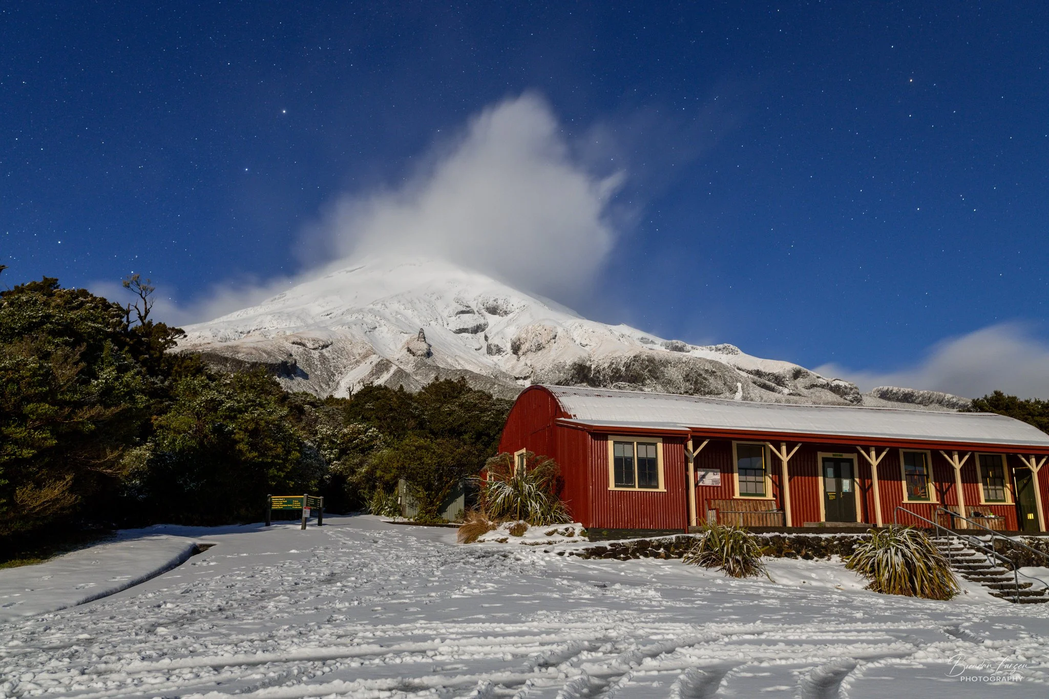 Snow-covered cabin in foreground with a snow-capped mountain and cloud in the background on a clear night sky filled with stars.