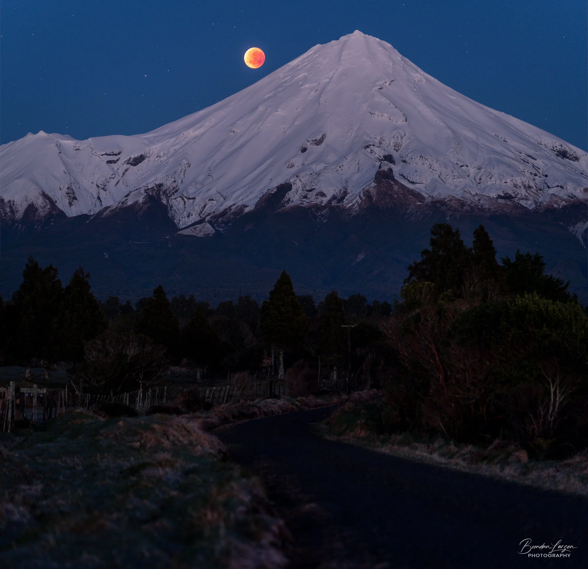Snow-capped mountain with a reddish moon in the sky, dark trees and a winding road in the foreground.