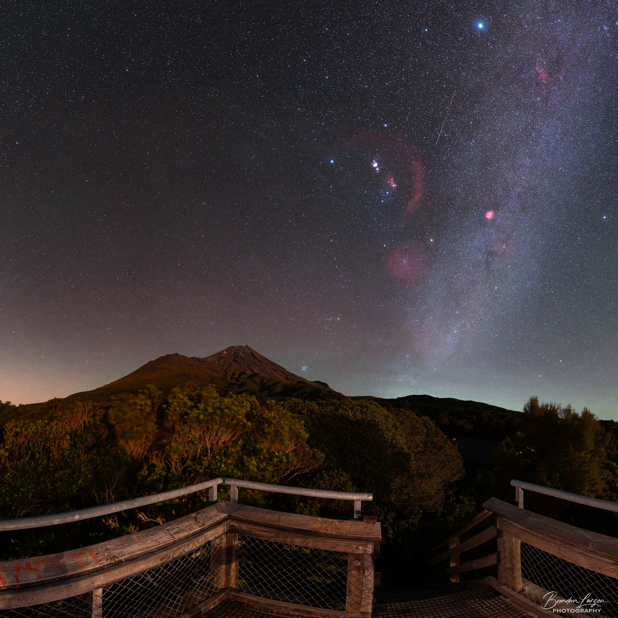 Night sky filled with stars and the Milky Way galaxy above a mountain and a wooden observation deck.