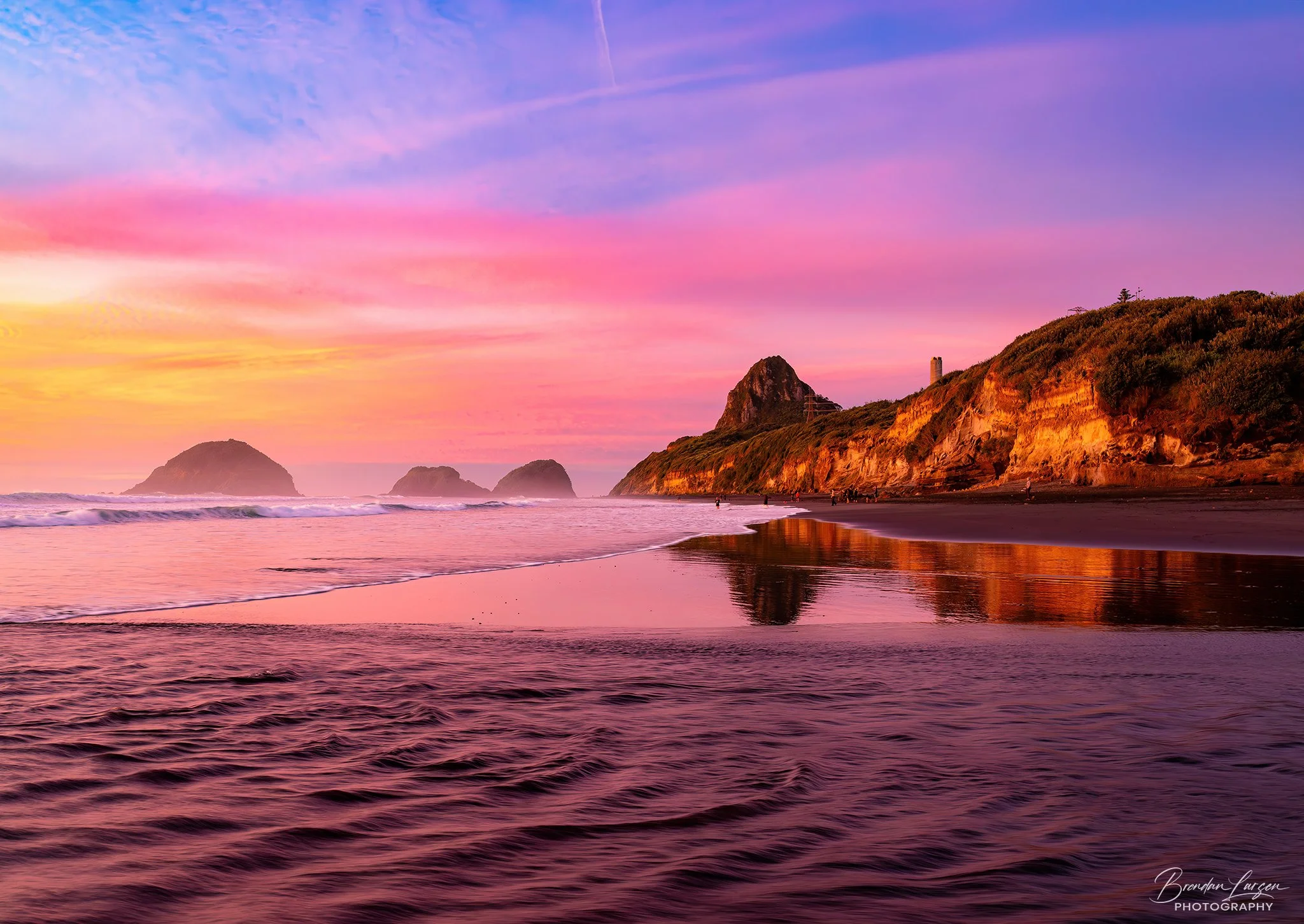 Sunset at a beach with pink, purple, and blue sky, cliffs on the right, waves and reflections on the shoreline, and distant rocks in the water.