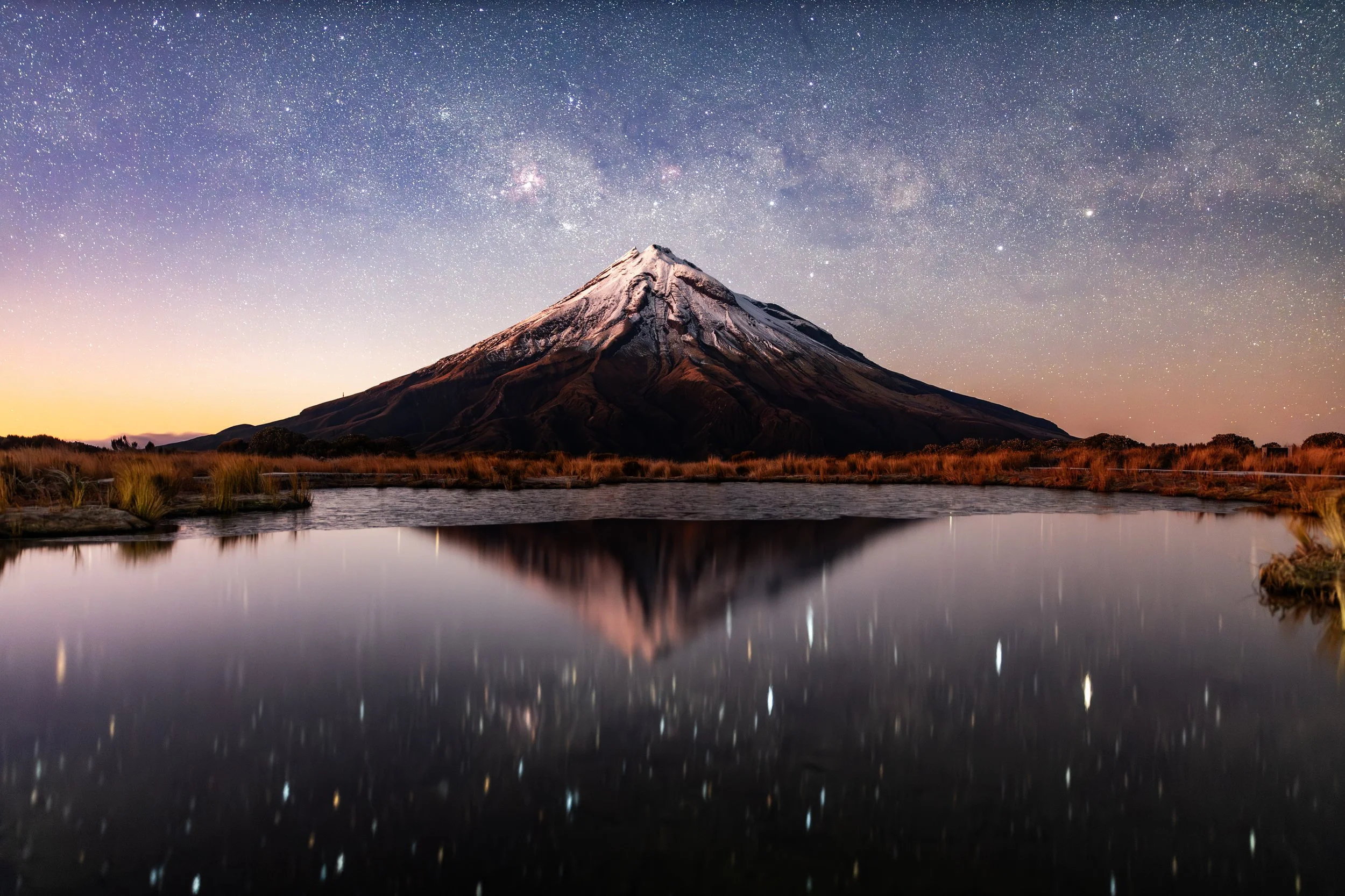 Night sky filled with stars over a snow-capped mountain reflected in a calm body of water.