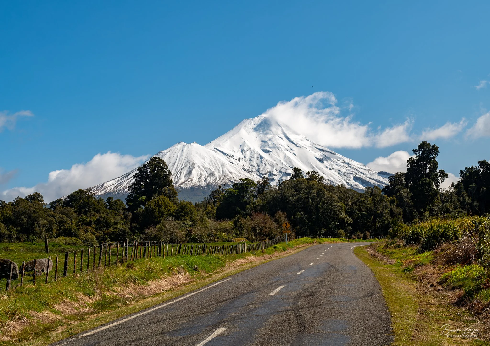 A winding country road leading toward snow-capped Mount Taranaki under a blue sky with white clouds.