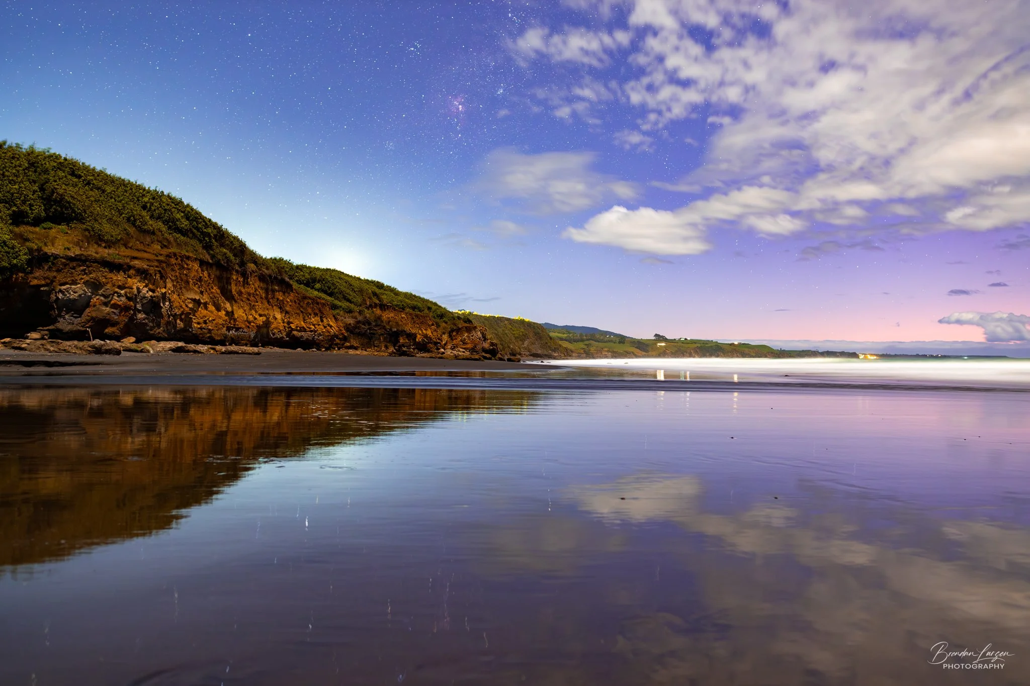 A night-time coastal scene with a starry sky, partly cloudy clouds, a rugged cliffside with greenery, and calm water reflecting the sky and landscape.
