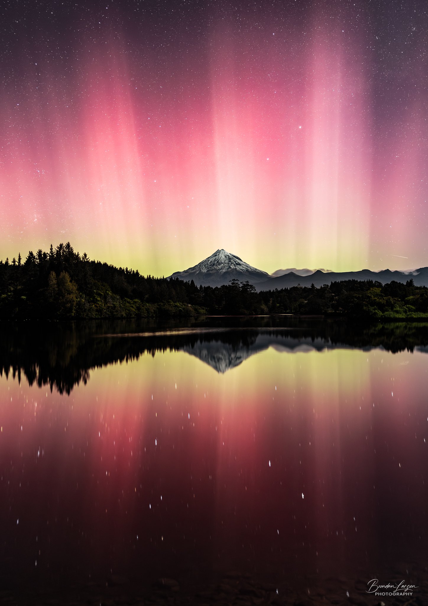 A vibrant display of the northern lights over a snow-capped mountain, with a calm lake reflecting the colorful sky and silhouette of trees.