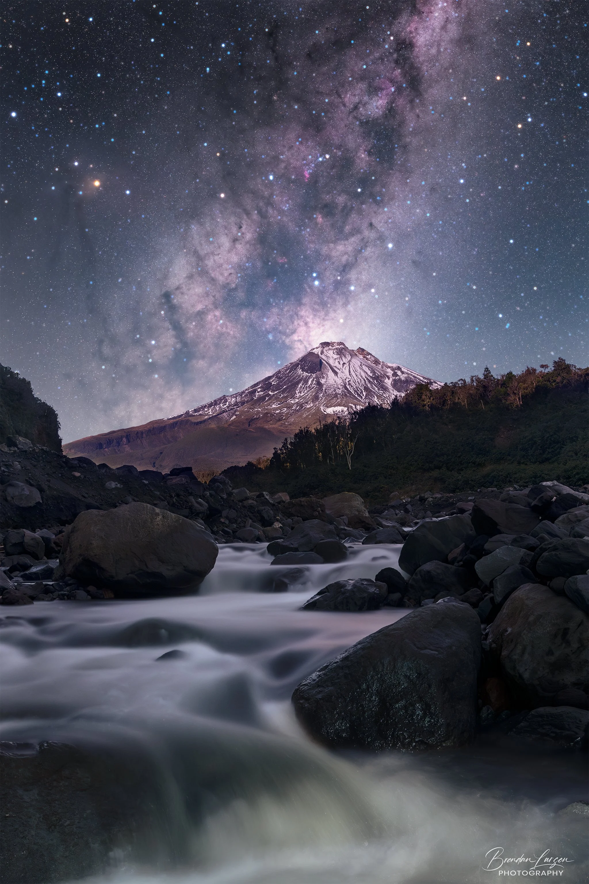 Nighttime scene of flowing river rocks with a snow-capped mountain under a starry sky, featuring the Milky Way galaxy.