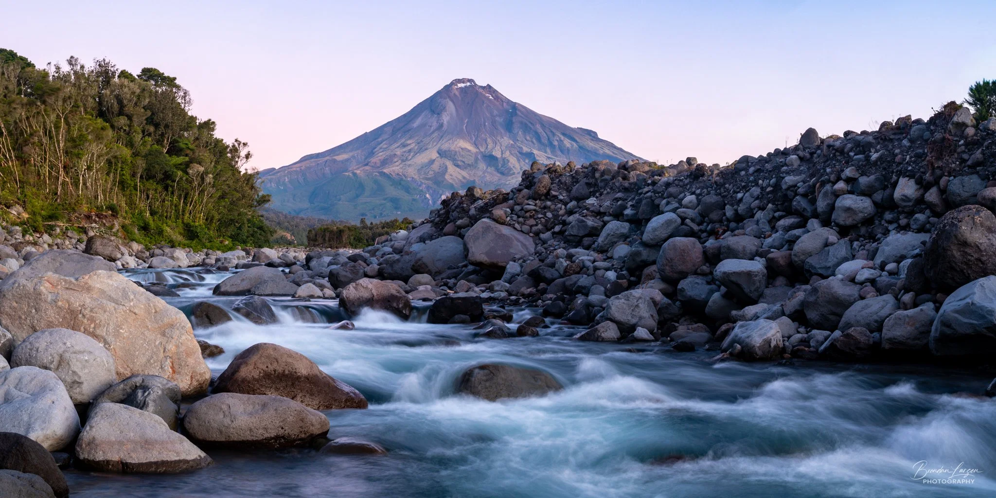 A mountain in the background, with a rocky river flowing in the foreground and green trees on the left side.