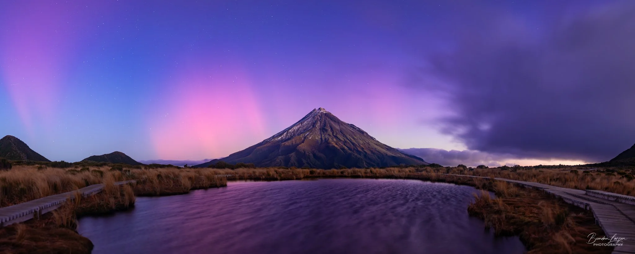 A scenic landscape featuring a tall mountain with a snow-capped peak, a calm water body reflecting the mountain, and a purple and pink aurora sky with clouds and stars.
