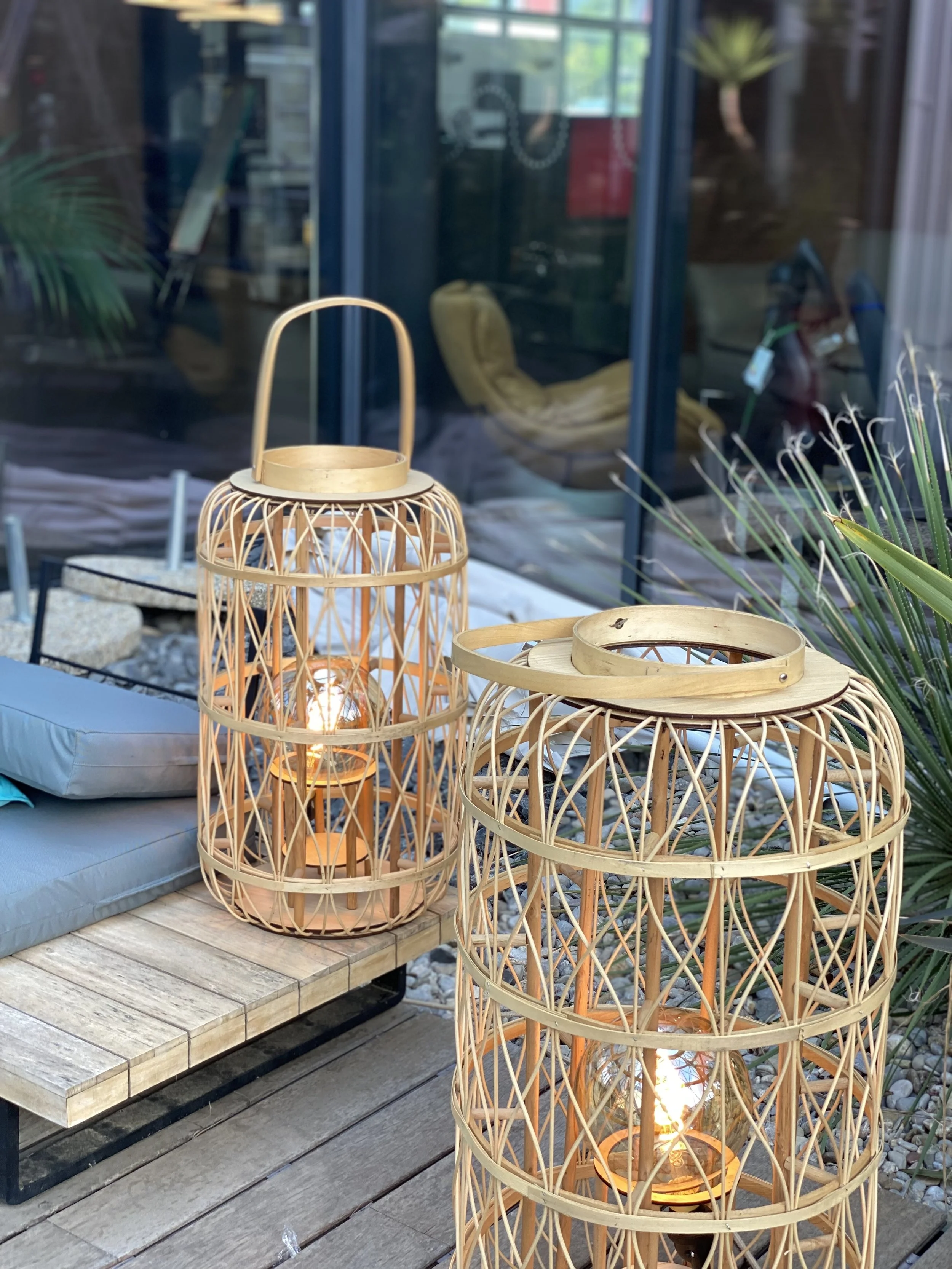 Deux lanternes en osier sur une terrasse en bois avec un canapé et des plantes en arrière-plan.