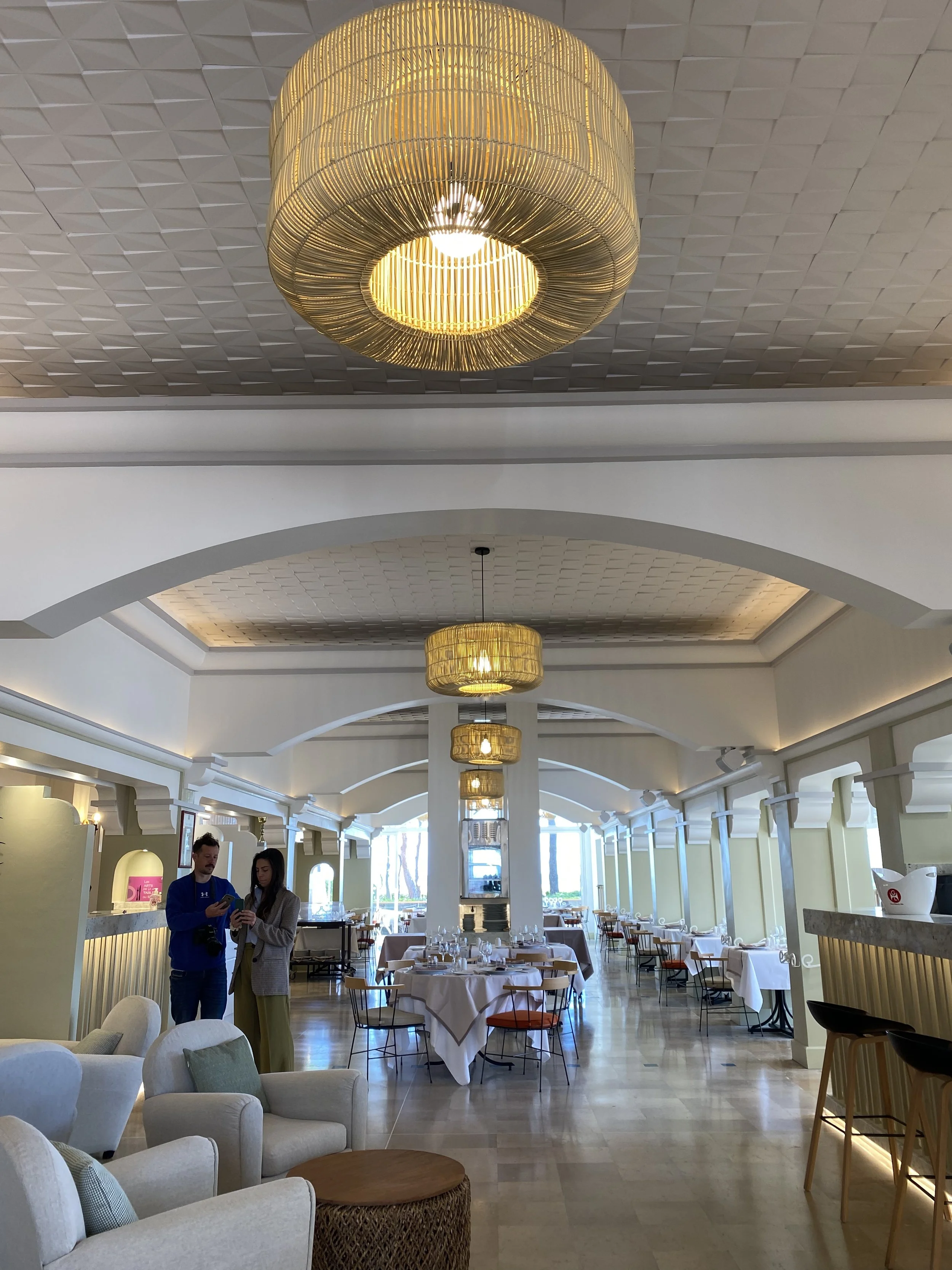 Intérieur lumineux d'un restaurant élégant avec des tables dressées et des suspensions modernes. Deux personnes discutent à proximité de la entrée, avec des fauteuils et tabourets au premier plan. Grandes baies vitrées en arrière-plan.