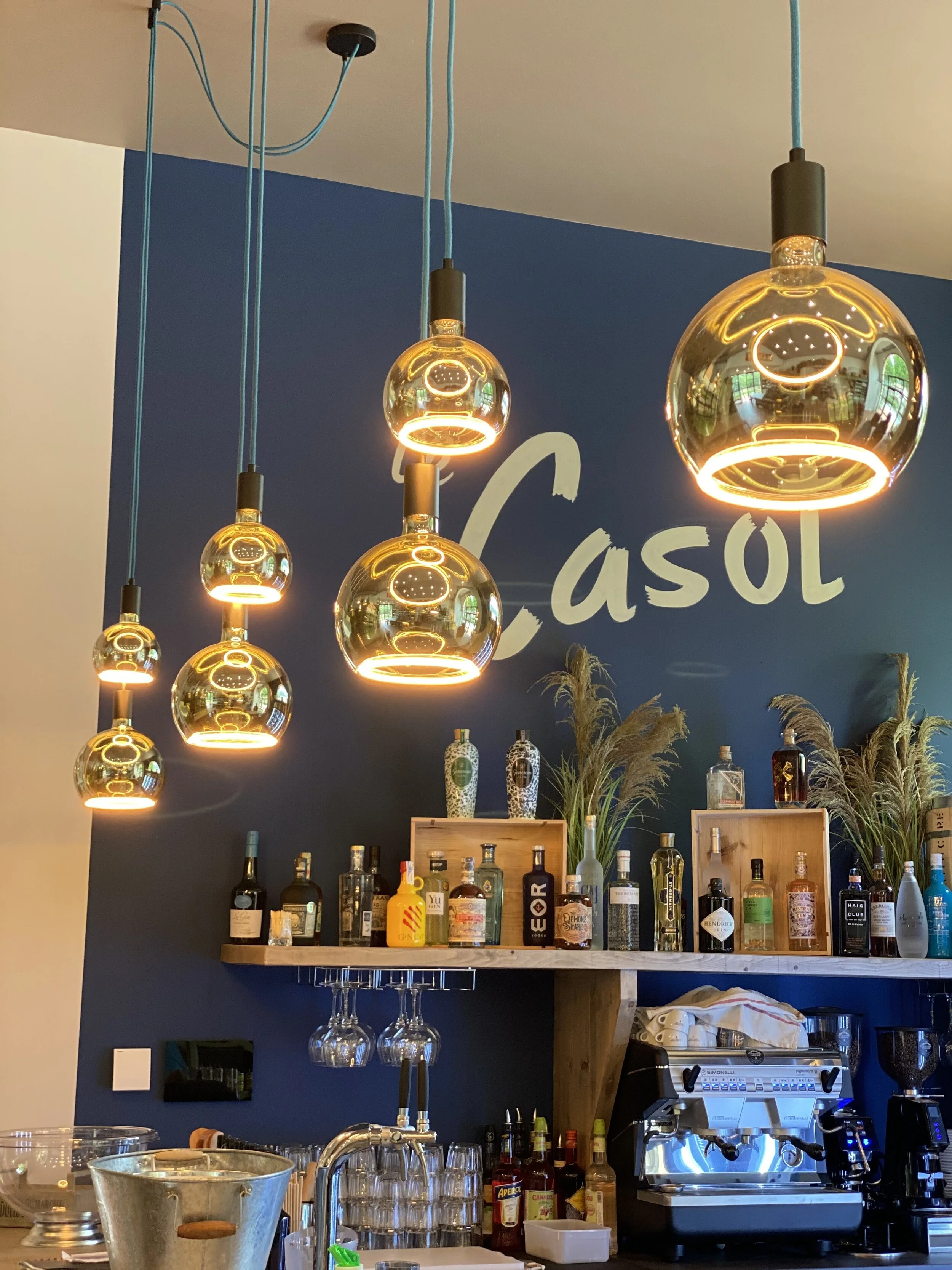 Intérieur d'un bar avec des luminaires suspendus modernes, des bouteilles d'alcool sur une étagère, des verres accrochés, une machine à café et une décoration murale bleue.