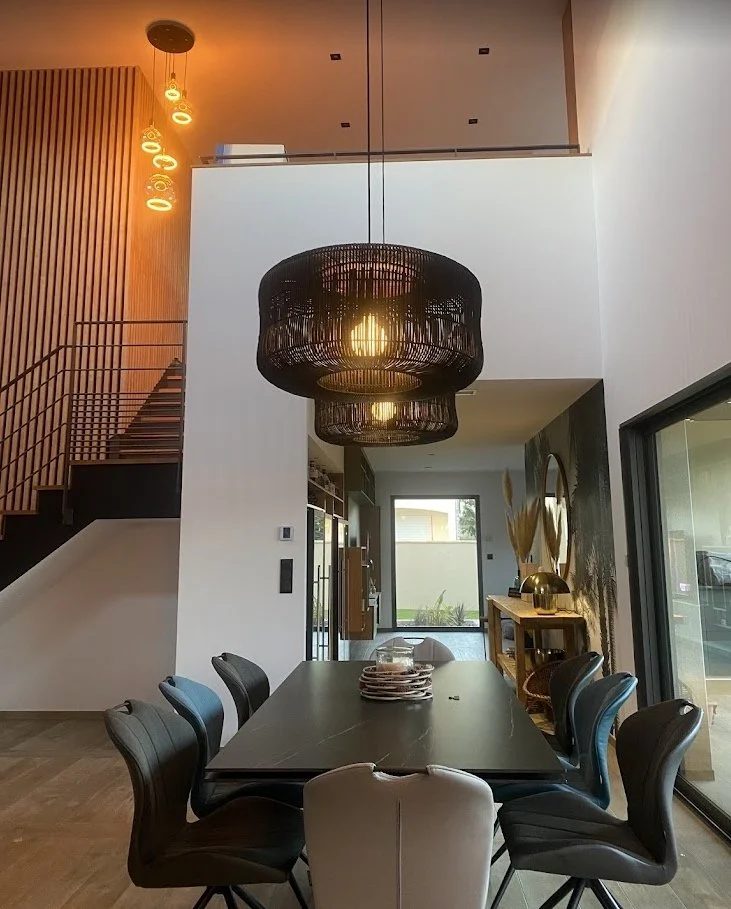 Salle à manger moderne avec table noire et chaises rembourrées, grand lustre suspendu, escalier en arrière-plan, et décoration intérieure élégante avec plantes et lumière naturelle.