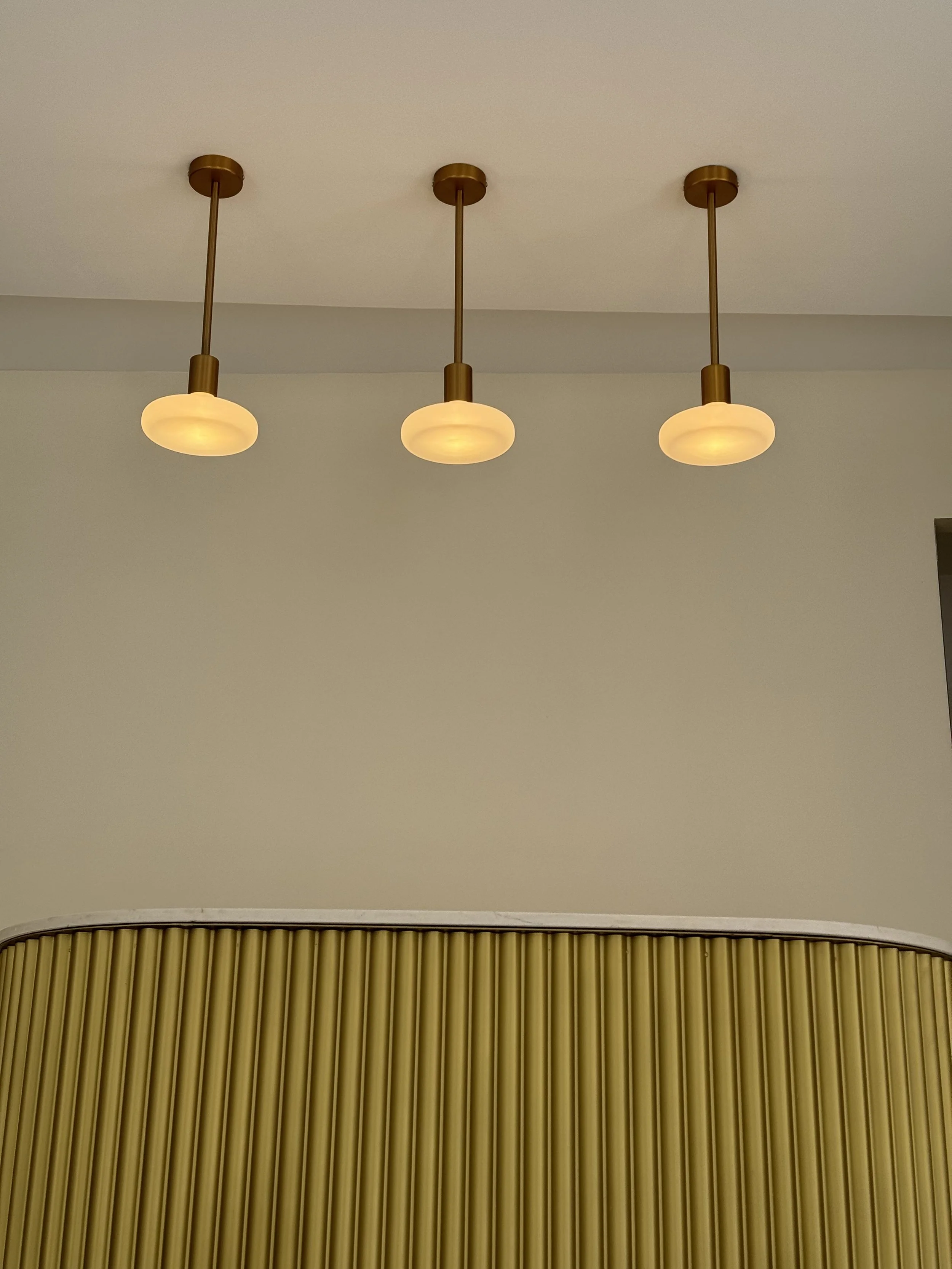 Trois suspensions lumineuses modernes accrochées au plafond au-dessus d'une cloison ondulée de couleur beige.