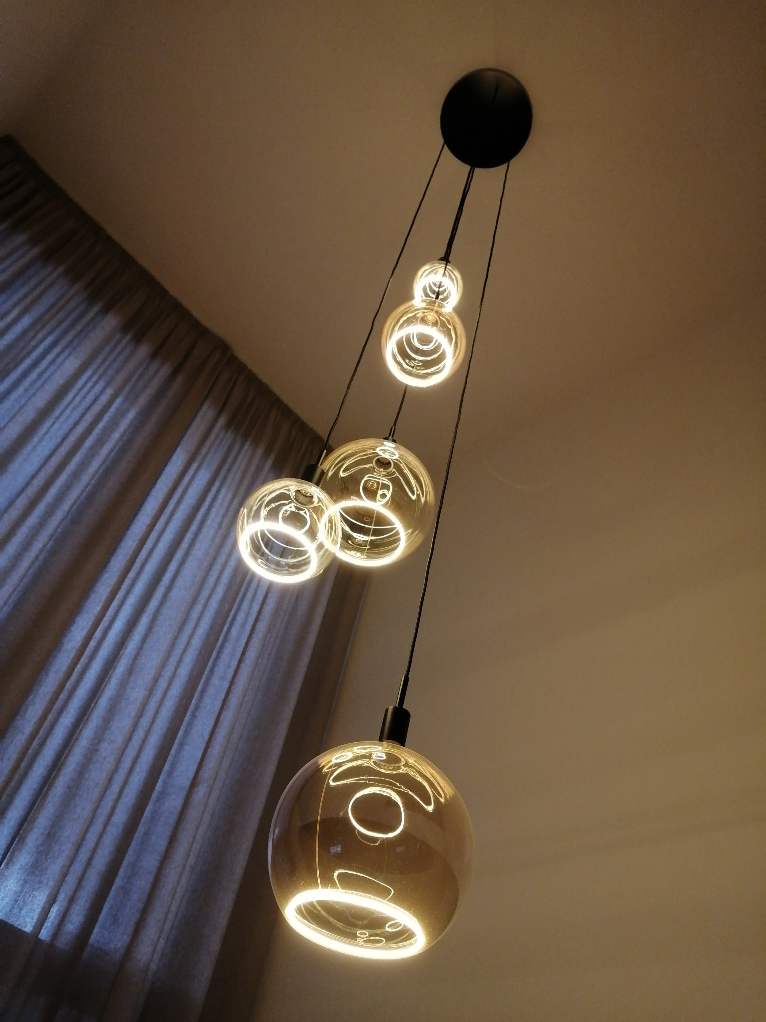 Lustre moderne avec plusieurs ampoules sphériques illuminées suspendues. Rideau à gauche.