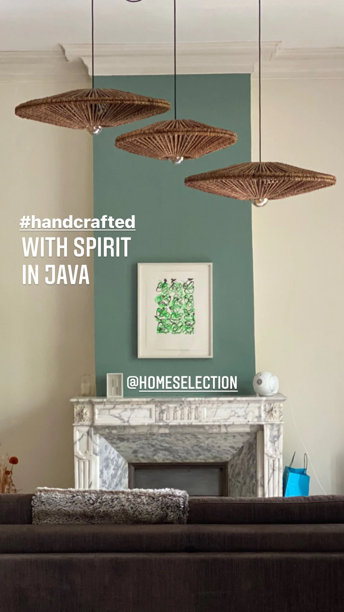 Salon avec trois suspensions en osier, une cheminée en marbre, un mur peint en vert avec un tableau dessus. Texte affiché : "#handcrafted with spirit in Java" et "@homeselection".