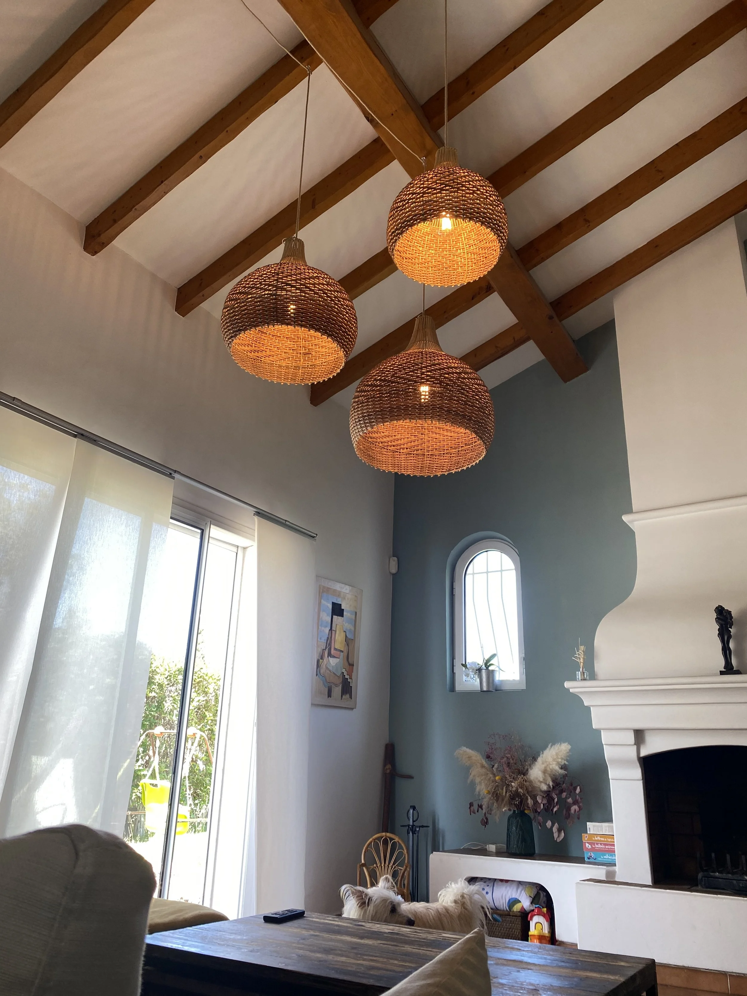 Intérieur d'un salon avec plafond en poutres apparentes, trois suspensions lumineuses en osier, une cheminée blanche, des rideaux clairs, et un décor avec des plantes et des livres.