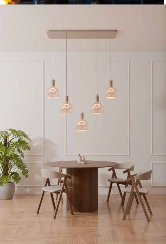 Une salle à manger moderne avec une table ronde en bois, quatre chaises en bois avec coussins, un luminaire suspendu avec cinq lampes en verre, et une grande plante verte dans un pot à gauche.