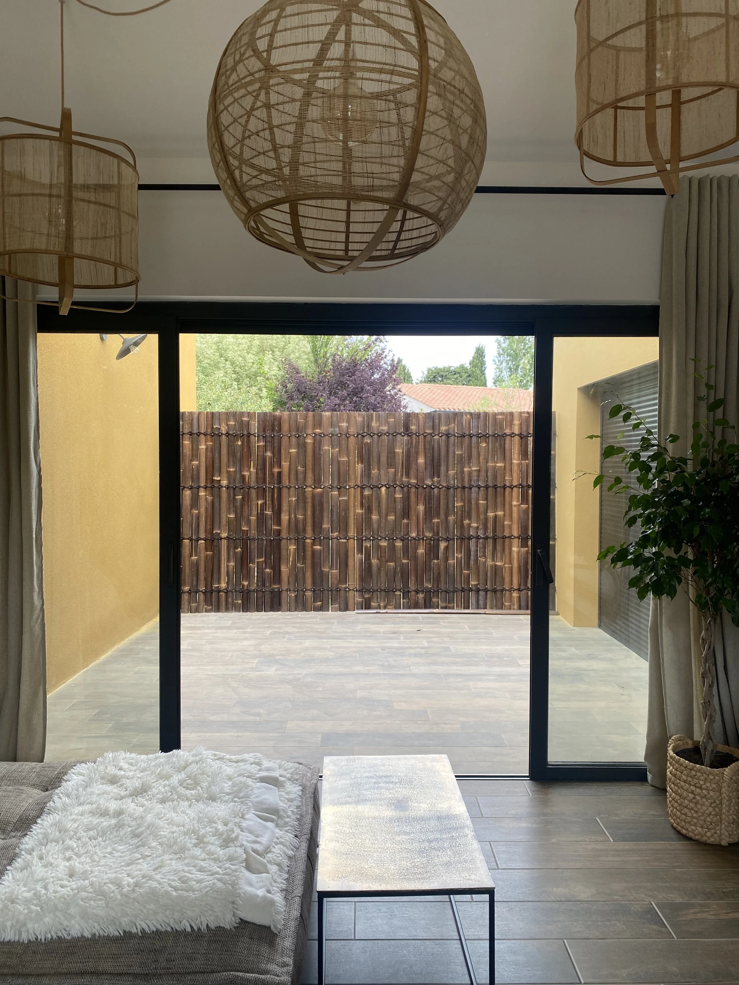 Intérieur moderne avec lampes suspendues en rotin, canapé avec couverture blanche, table basse et grande fenêtre donnant sur une cour avec clôture en bois.