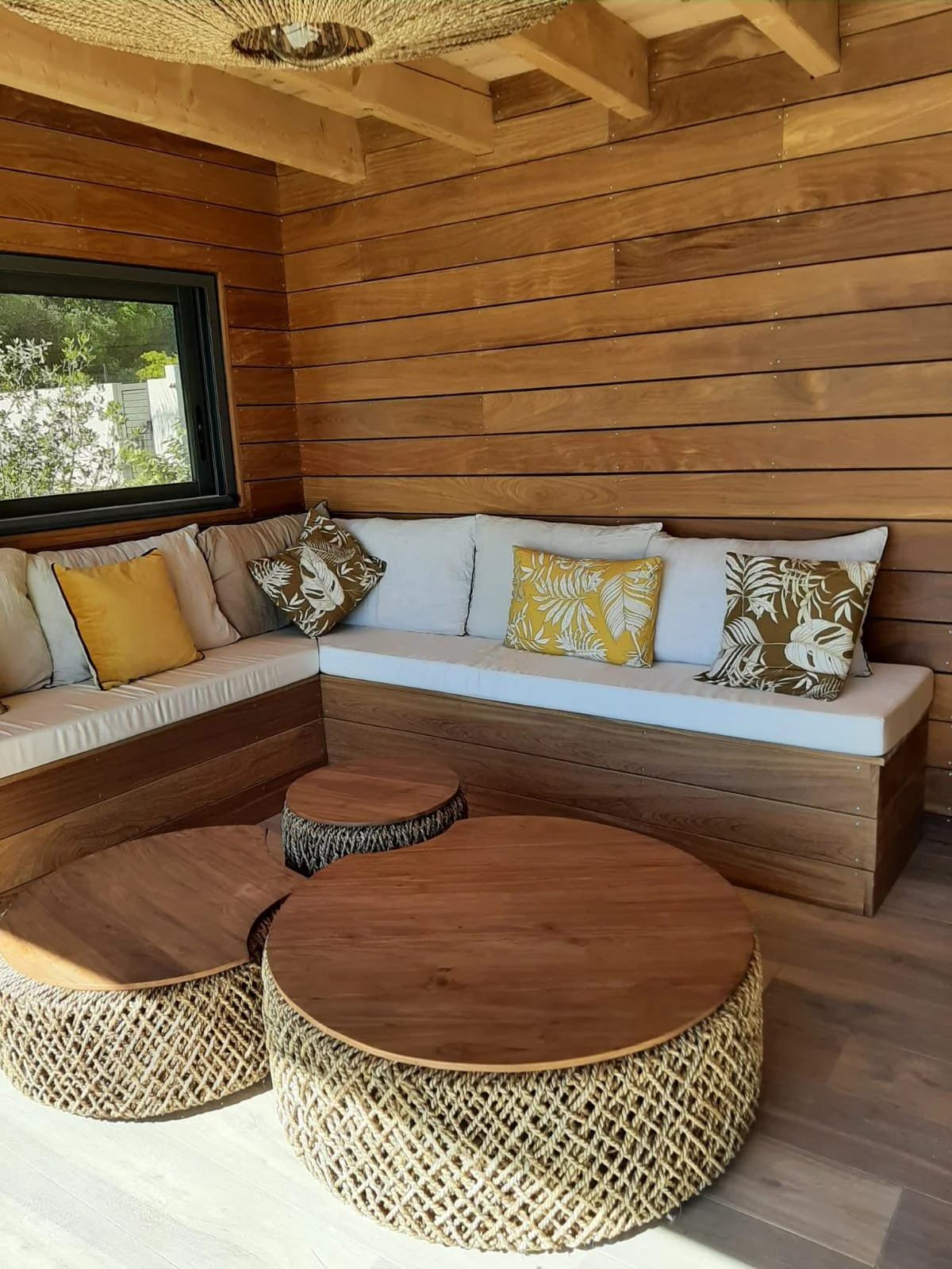 Terrasse en bois avec canapé d'angle, coussins décoratifs jaune et marron, et tables basses rondes en bois et osier.