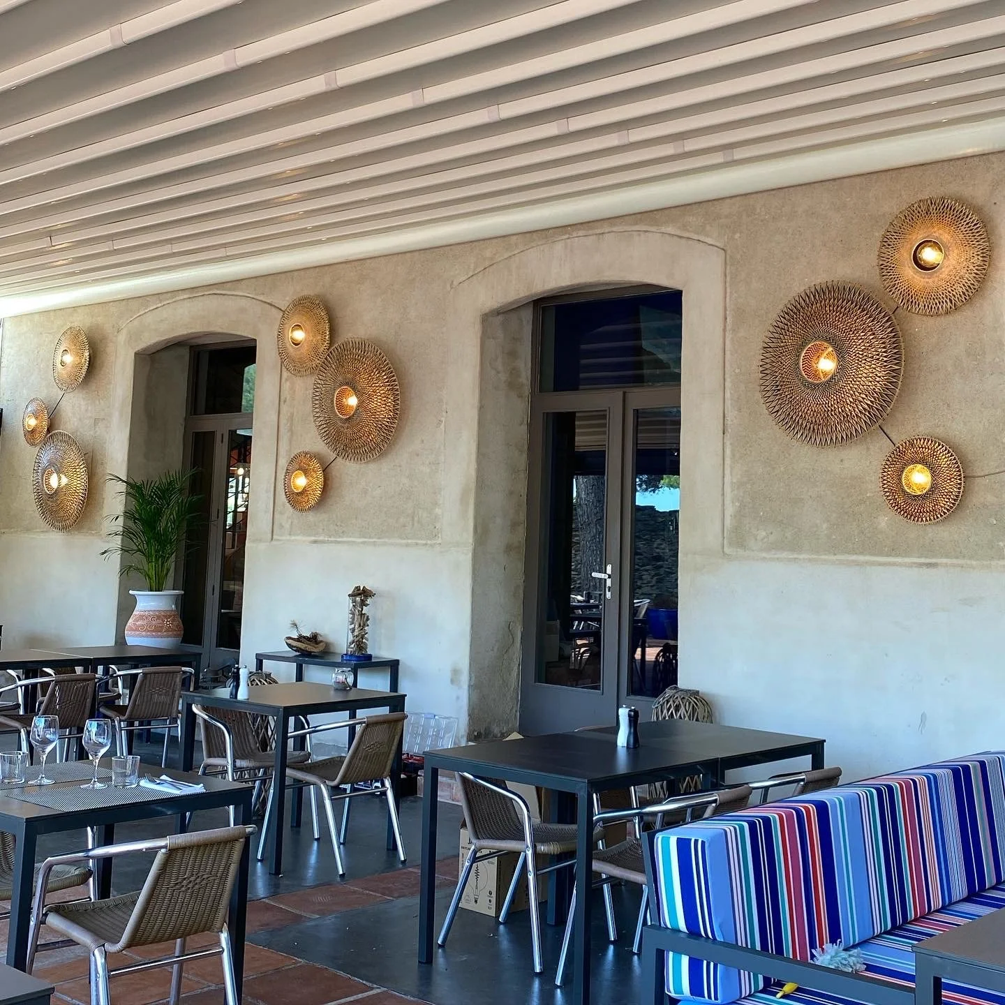 Intérieur d'un restaurant avec des tables et des chaises en osier, des décorations murales circulaires en rotin avec éclairage, et un canapé à rayures multicolores.