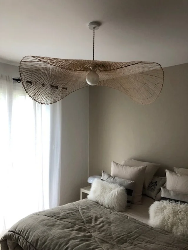 Chambre à coucher avec un grand lit double recouvert de coussins blancs et gris, éclairé par un plafonnier suspendu au design moderne en rotin. La pièce a un mur crème clair et une fenêtre avec un voile blanc laissant entrer la lumière du jour.