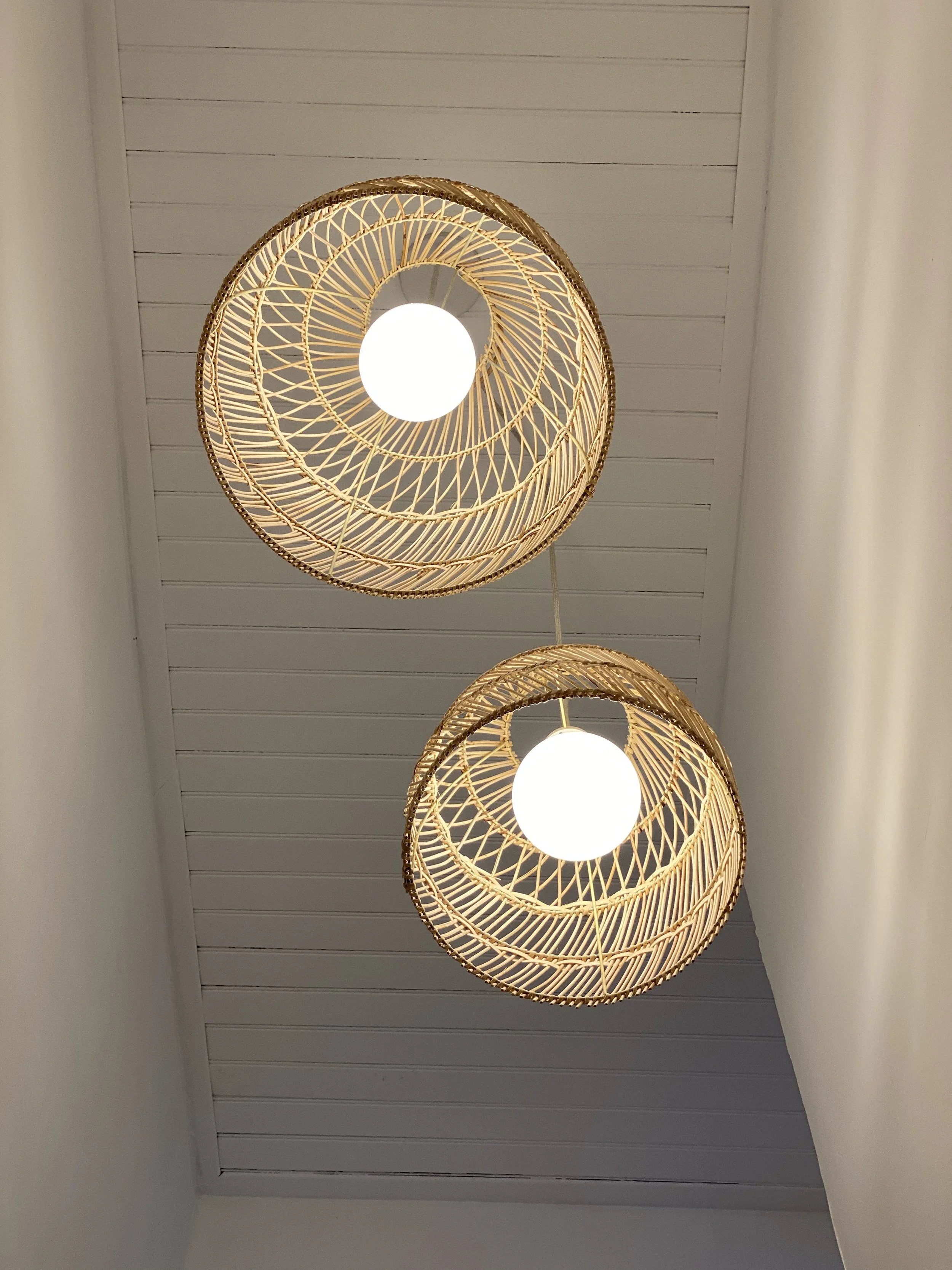 Deux suspensions en osier avec des ampoules allumées, fixées à un plafond en bois blanc.