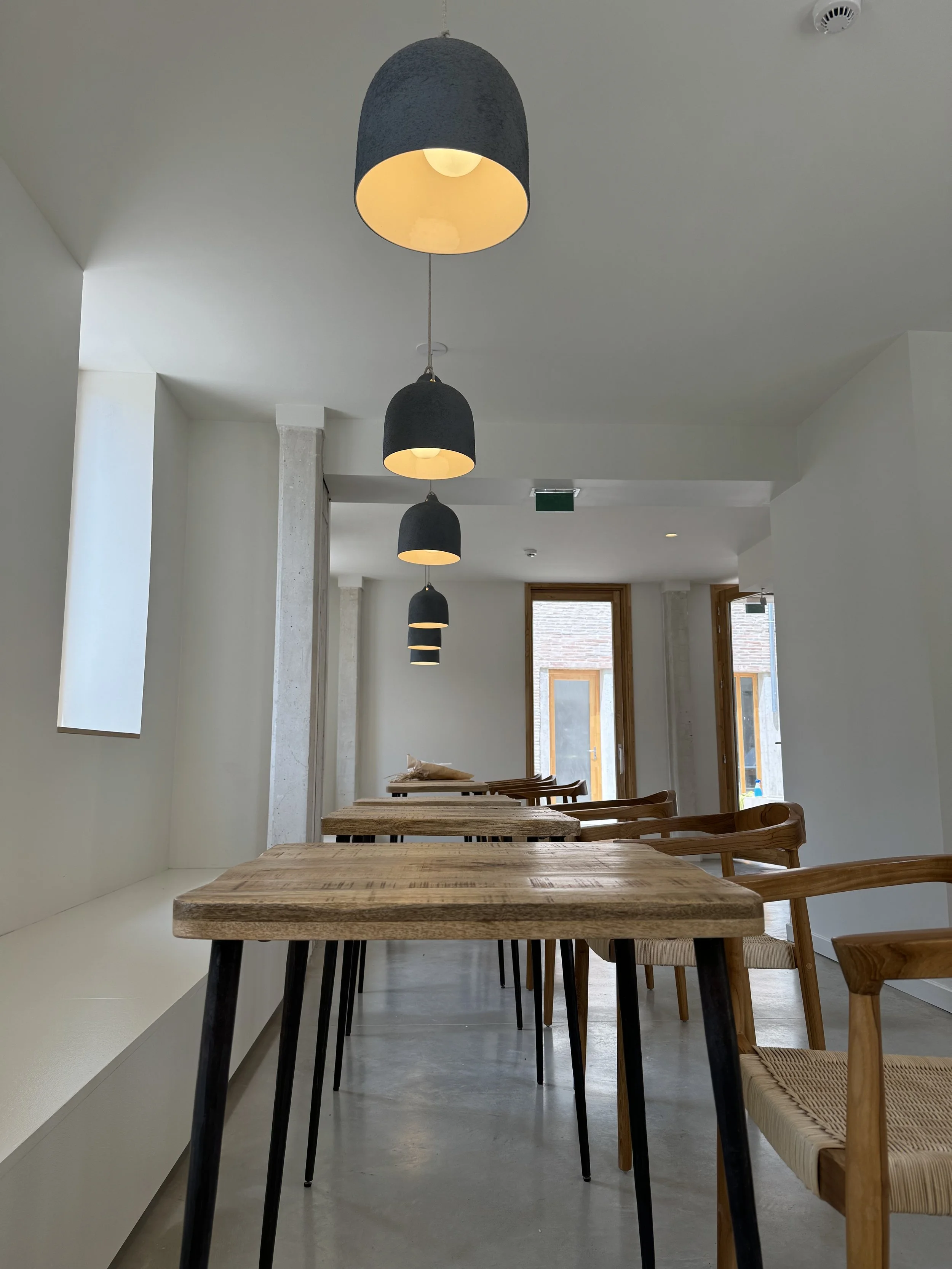 Intérieur d'un café moderne avec tables en bois et chaises en osier, lumières suspendues au plafond, et grandes fenêtres laissant entrer la lumière naturelle.