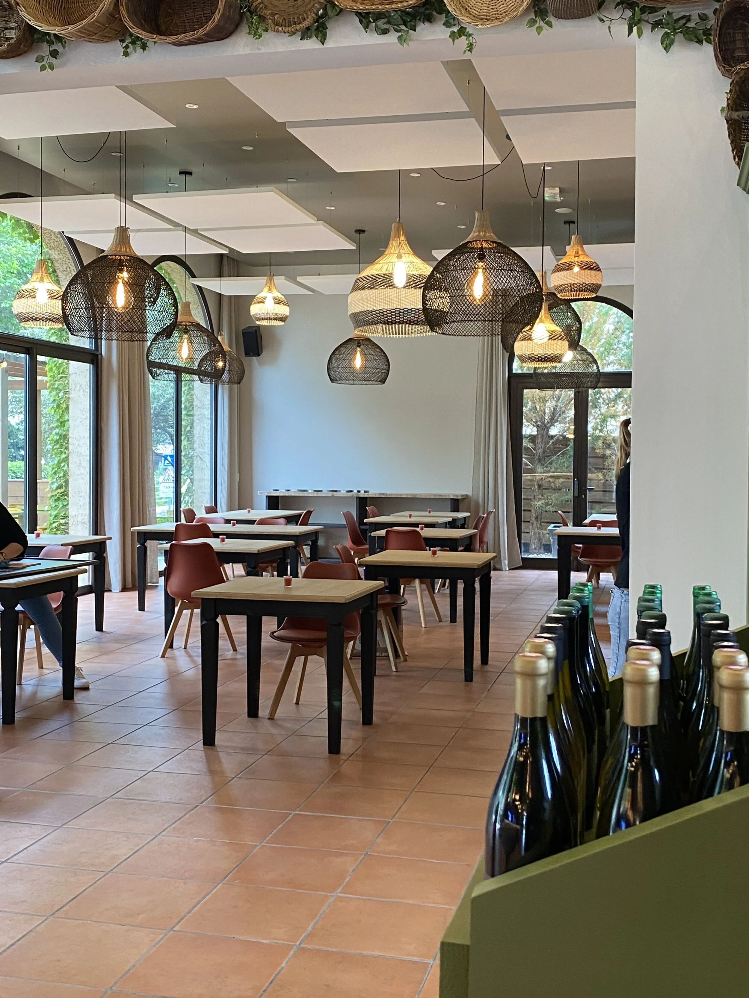 Salle de restaurant moderne avec des tables en bois, des chaises rouges, des luminaires suspendus et des bouteilles de vin en avant-plan.