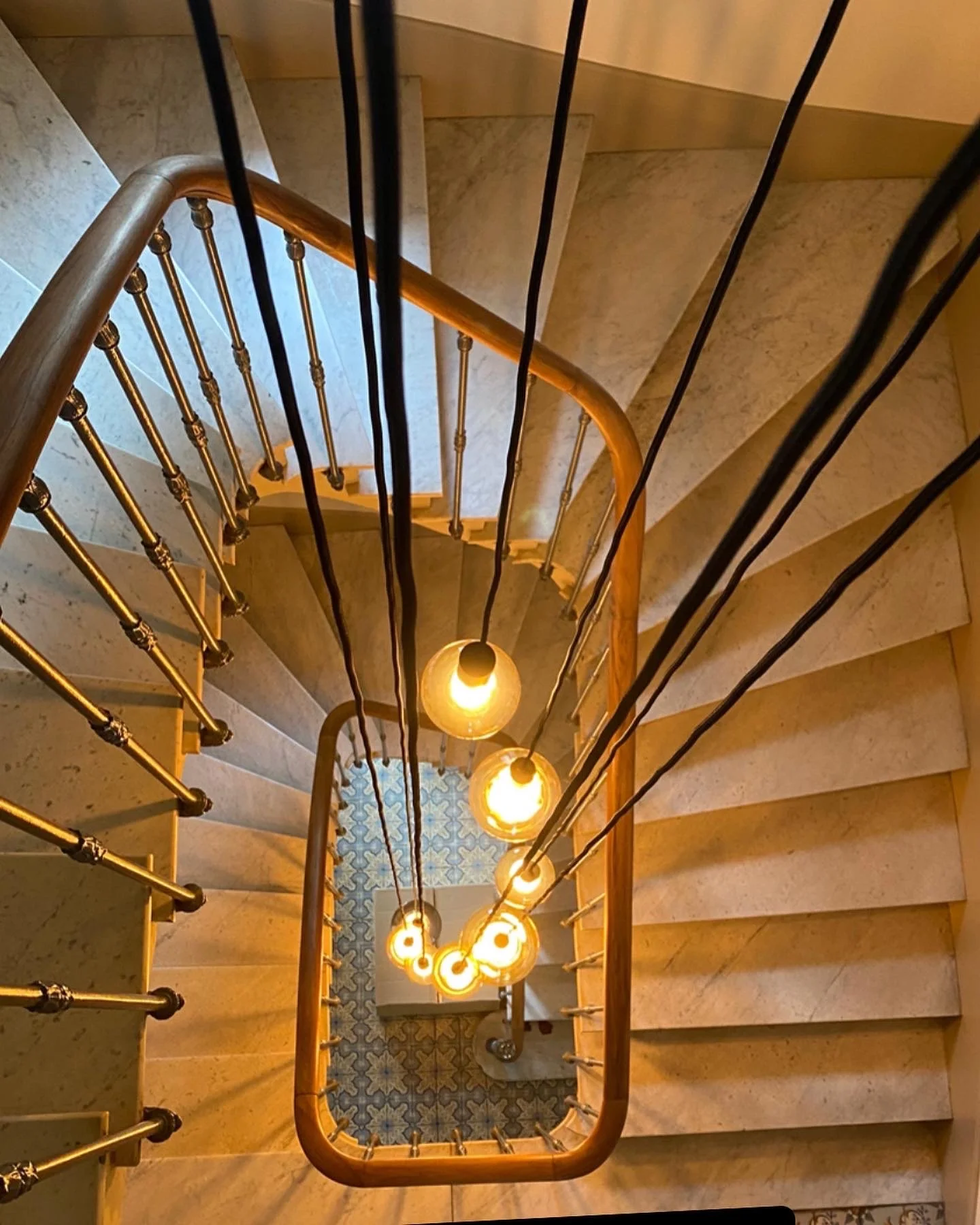 Vue plongeante d'un escalier en colimaçon avec balustrade en bois et éclairage suspendu au centre, éclairant les marches en marbre.
