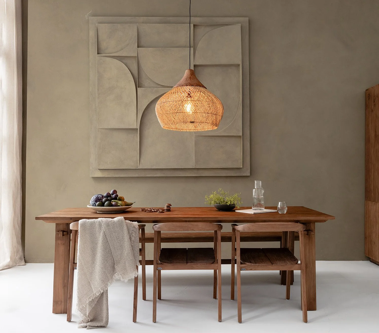 Salle à manger avec table en bois, chaises, lampe suspendue en osier, assiette de fruits, carafe d'eau et tableau moderne au mur.