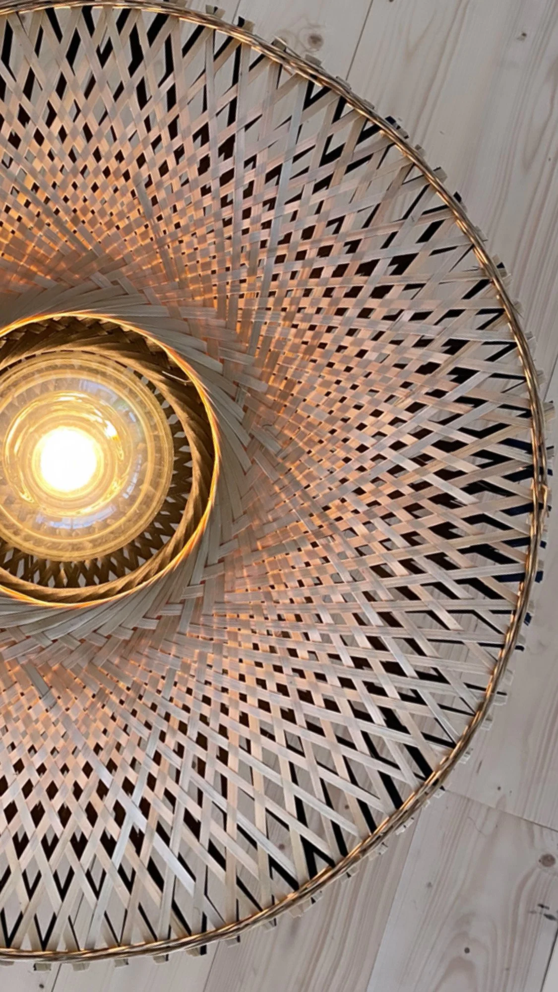 Vue de dessous d'une lampe suspendue avec un abat-jour en rotin tissé autour d'une ampoule allumée, plafond en bois en arrière-plan.
