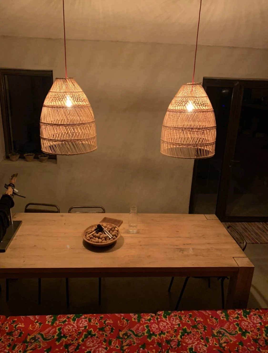 Intérieur avec deux suspensions en rotin au-dessus d'une table en bois, une carafe d'eau, un bol de noix, et des chaises. Décor sombre et cozy.