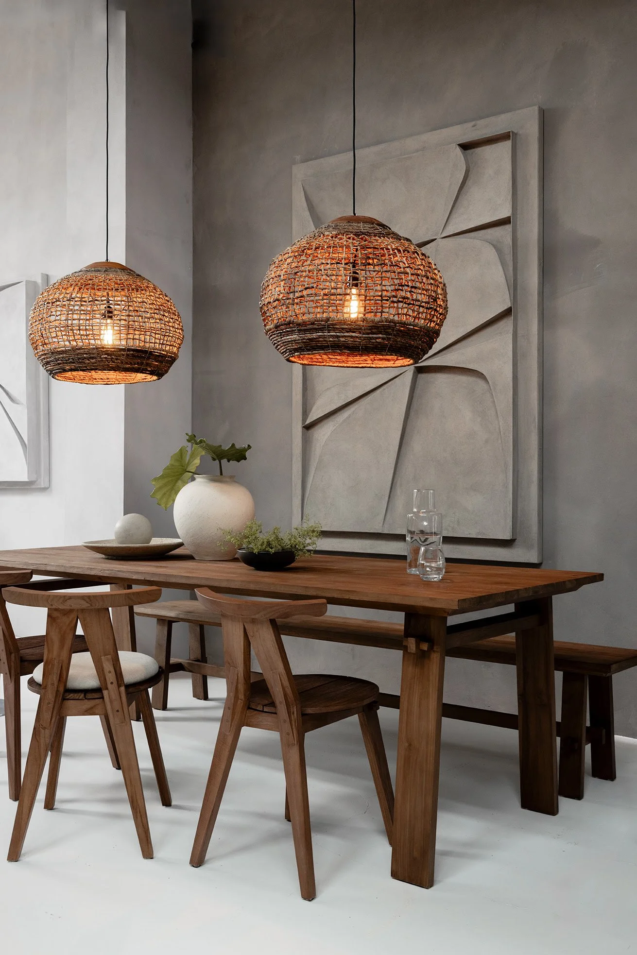 Salle à manger moderne avec table en bois, chaises assorties, lampes suspendues en rotin, décoration murale artistique et vase blanc sur la table.