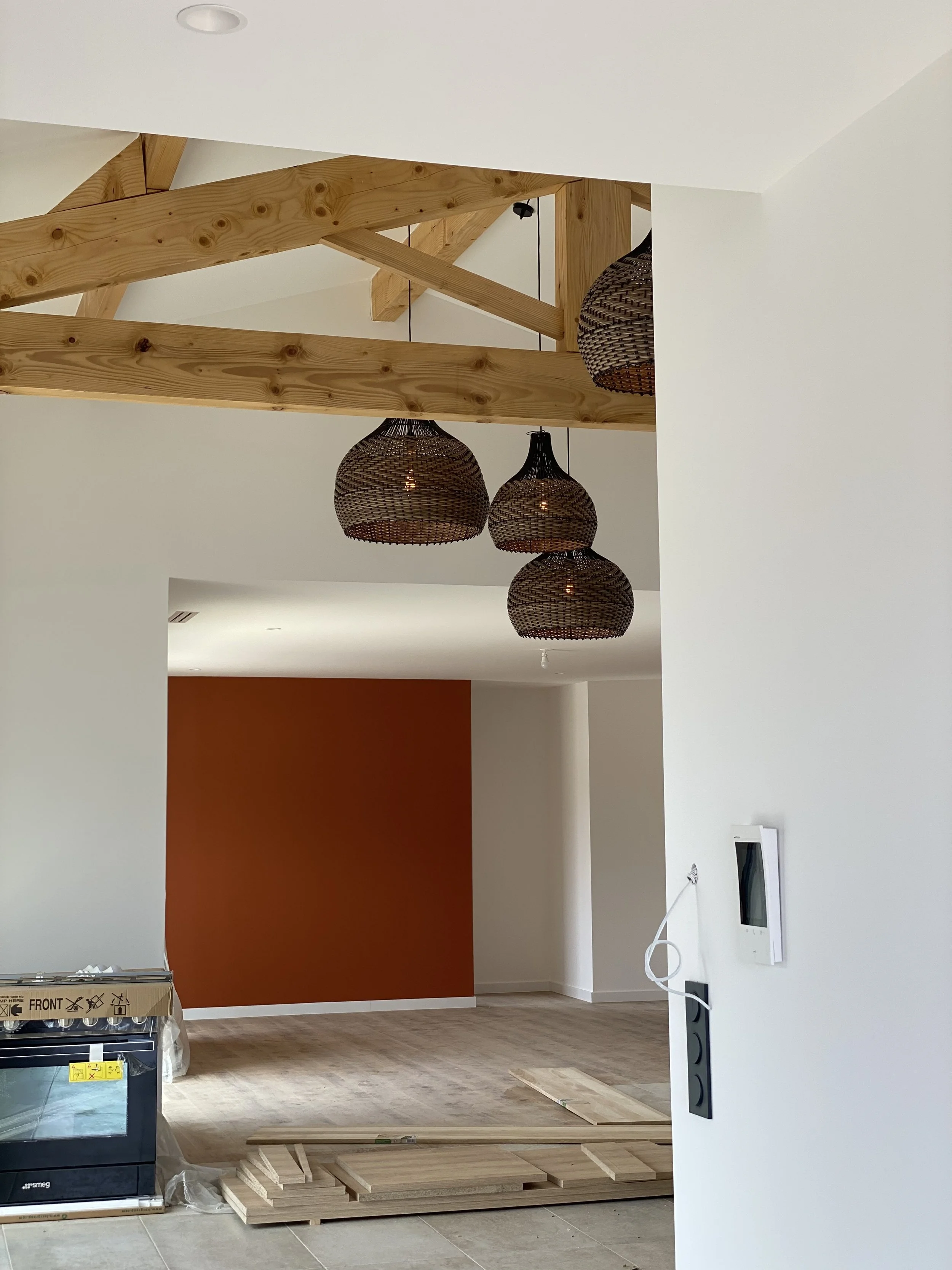 Intérieur d'une pièce en rénovation avec poutres apparentes en bois, suspensions en osier, mur orange, et matériel de construction au sol.
