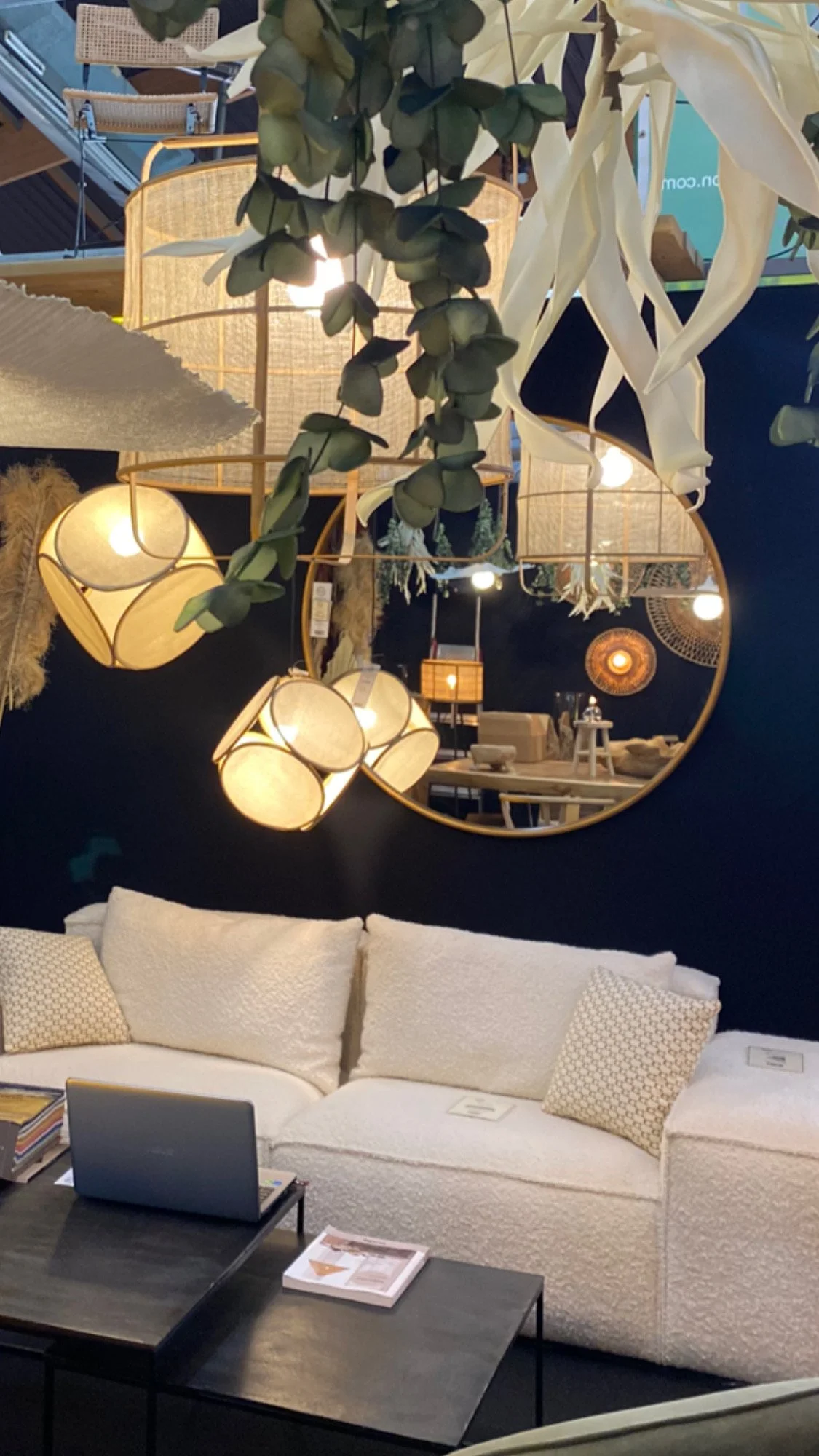 Salon moderne avec canapé blanc, table basse noire, ordinateur portable, miroir rond, plantes suspendues et lampes décoratives.
