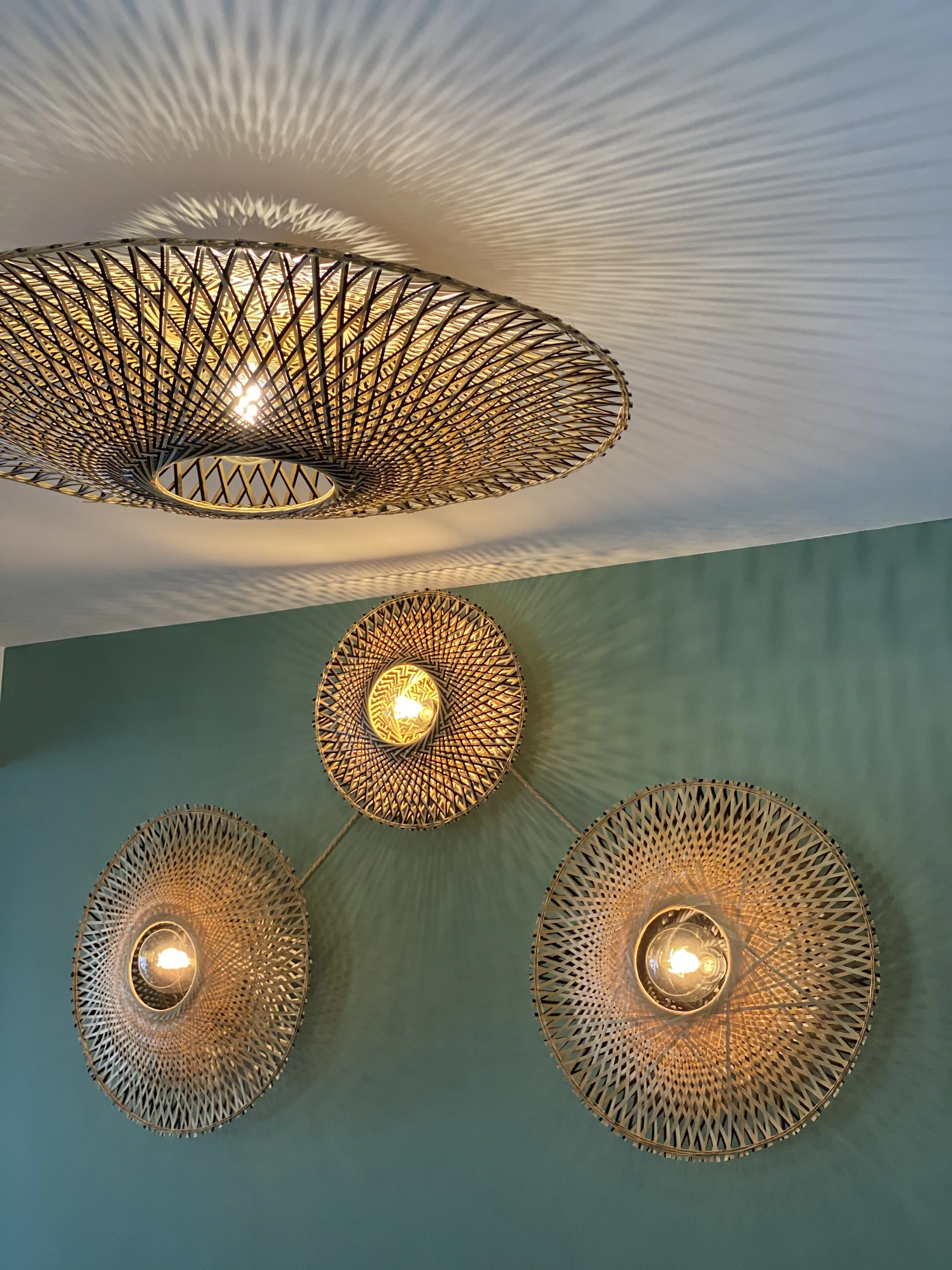 Luminaires en rotin tissés contre un mur vert et plafond blanc, produisant des motifs d'ombre complexes.