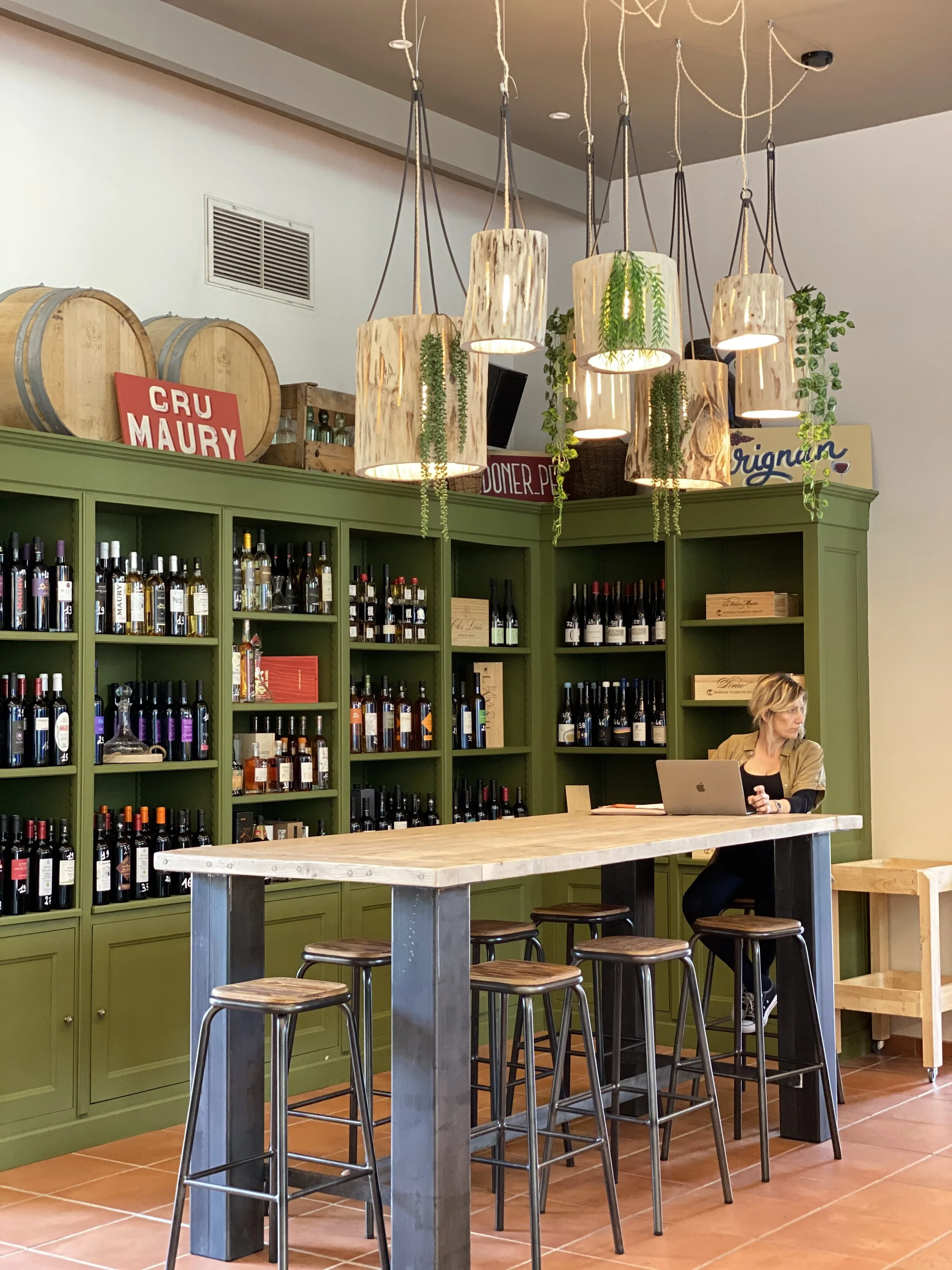Intérieur d'une cave à vin avec des étagères vertes remplies de bouteilles de vin, un bar en bois avec des tabourets, des tonneaux décoratifs, suspensions lumineuses avec des plantes, et une personne utilisant un ordinateur portable.