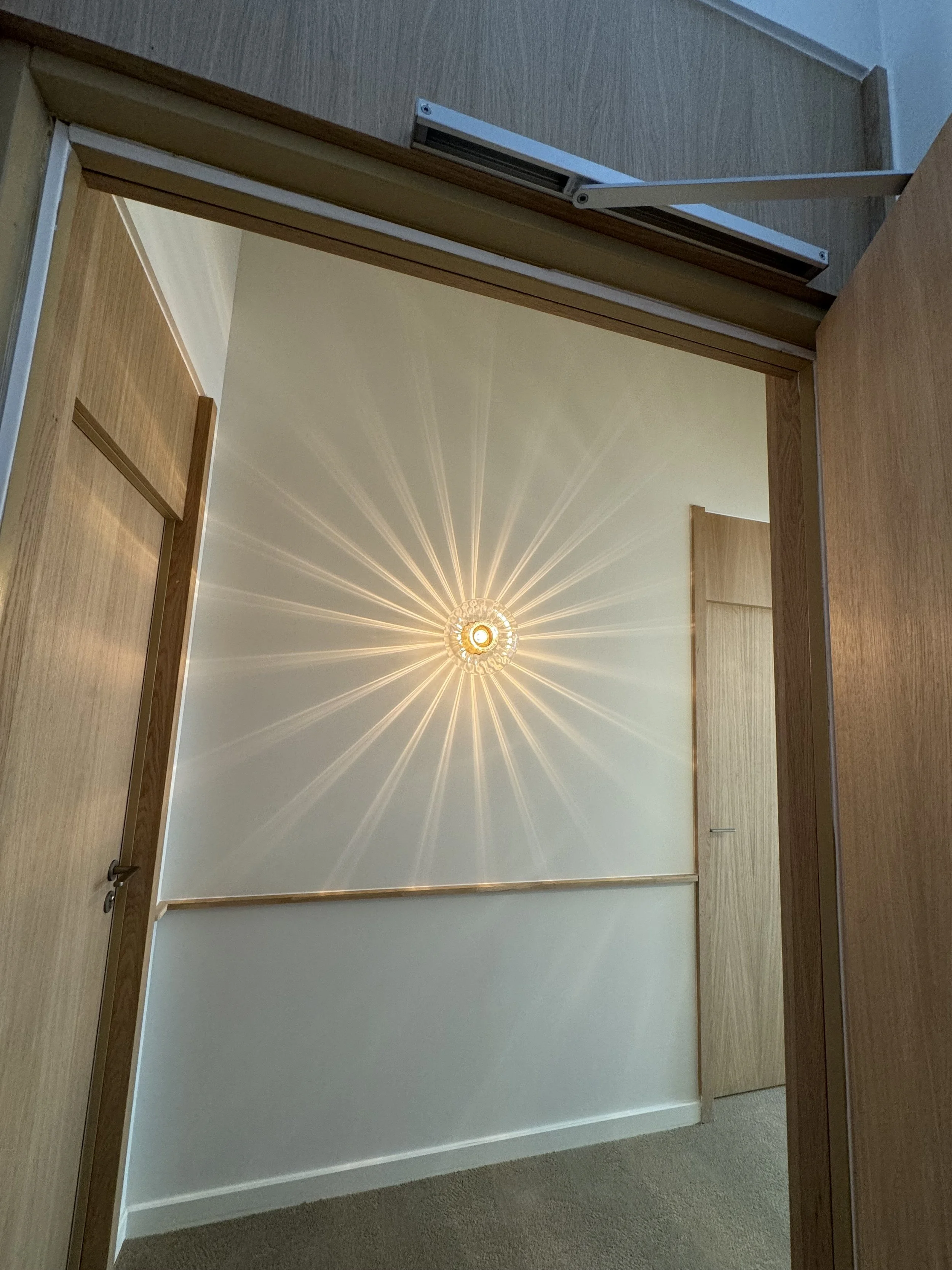 Vue d'un couloir avec une applique murale éclairée, émettant des rayons lumineux sur le mur, encadrée par des portes en bois clair.