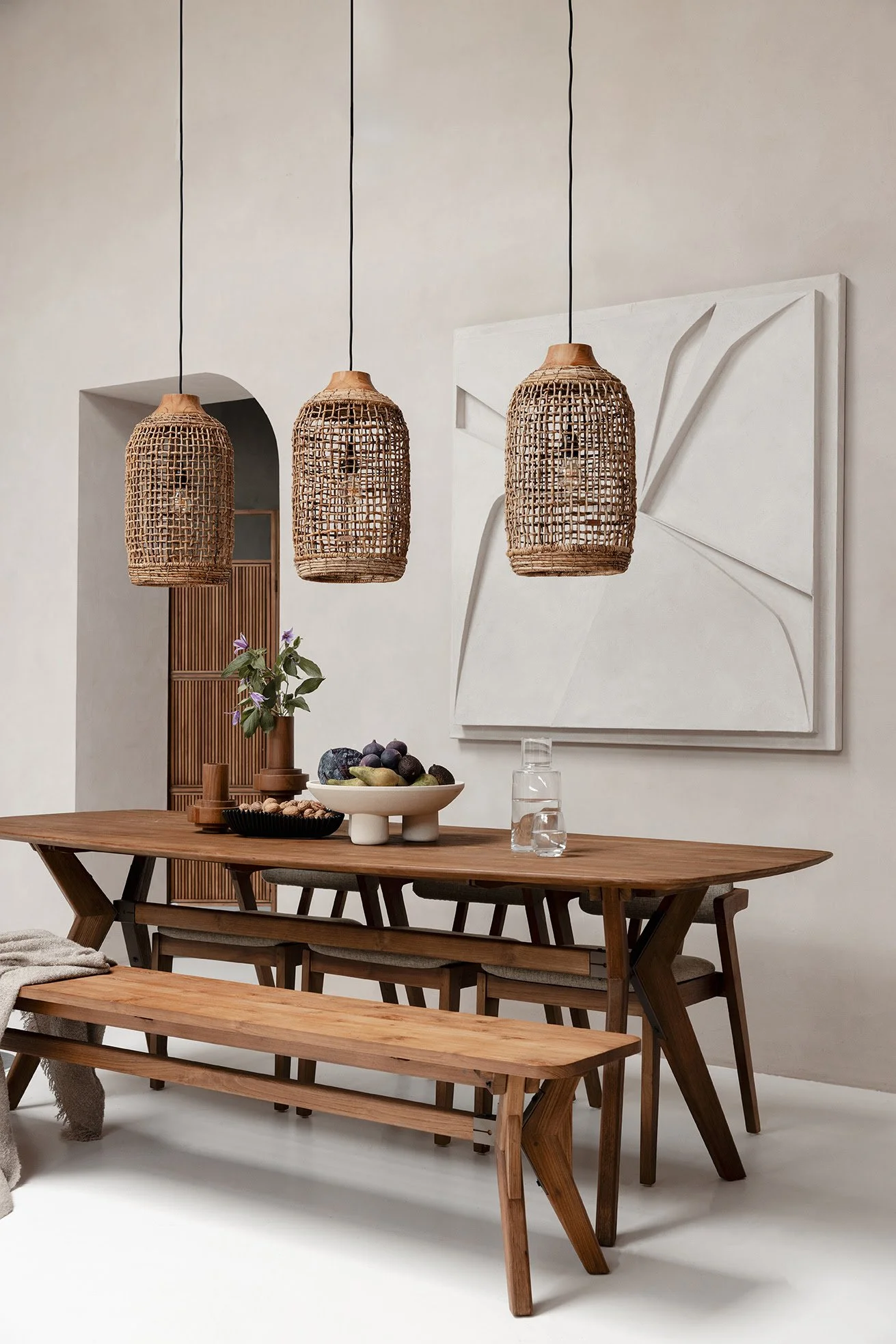Salle à manger avec table et bancs en bois, suspension en osier, tableau mural géométrique, décorations de fruits et plantes.
