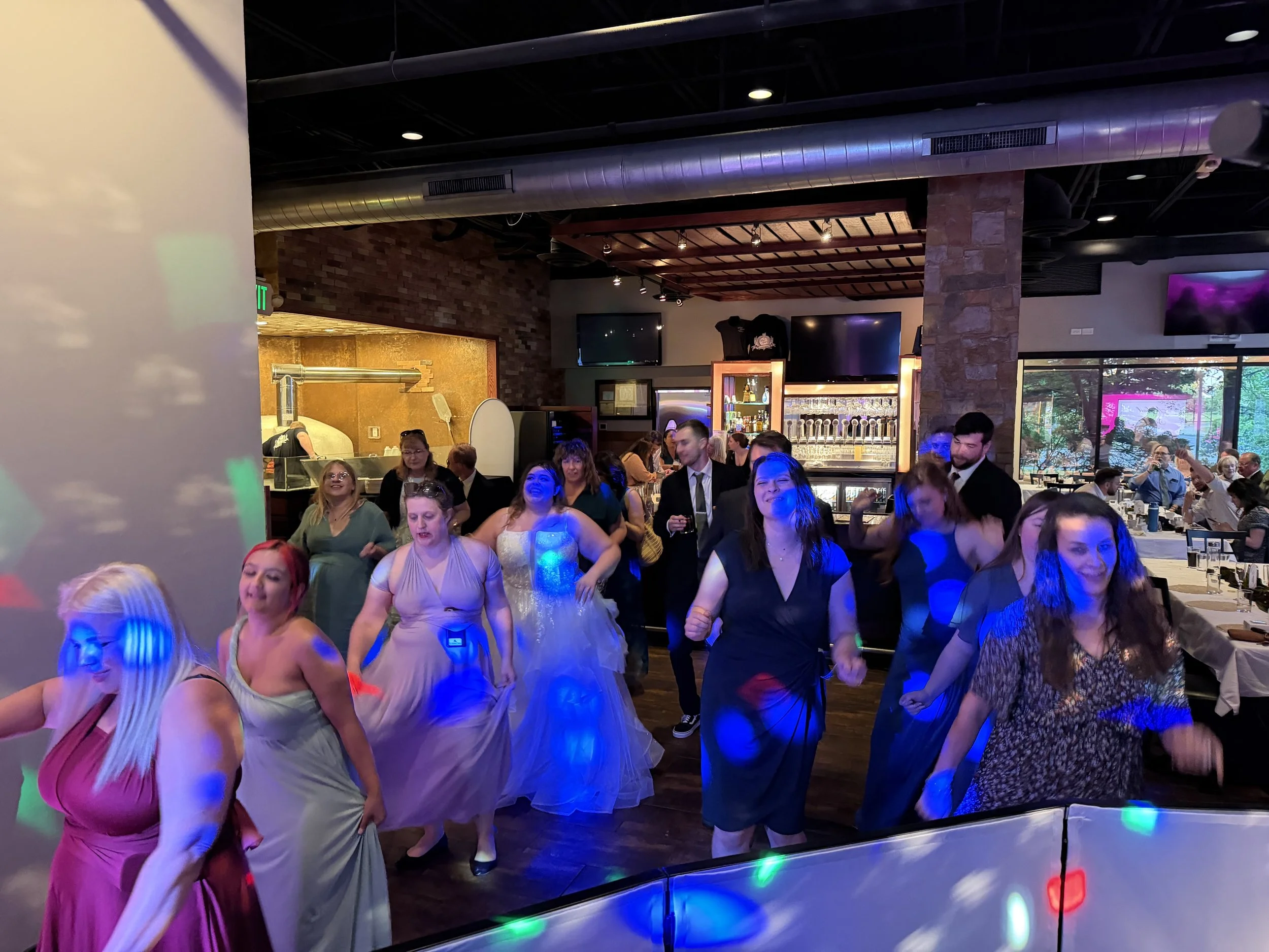 People dancing at a celebration inside a restaurant or event space with exposed brick walls and a bar area in the background.