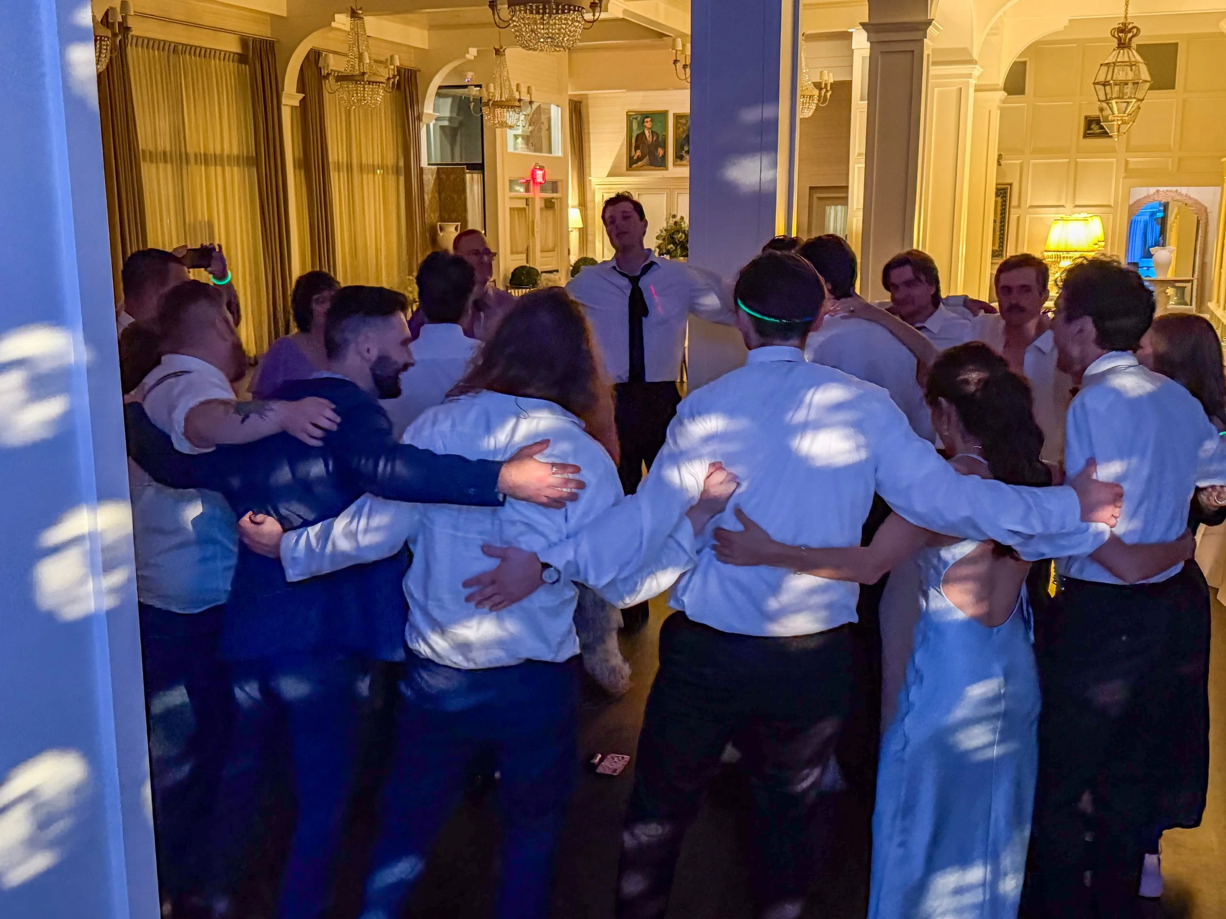 A group of people gathered in a warmly lit, elegant room, hugging and celebrating. They are dressed in formal or semi-formal attire, with some wearing white shirts. The room features chandeliers, paintings, and a yellow lamp, indicating a sophisticat