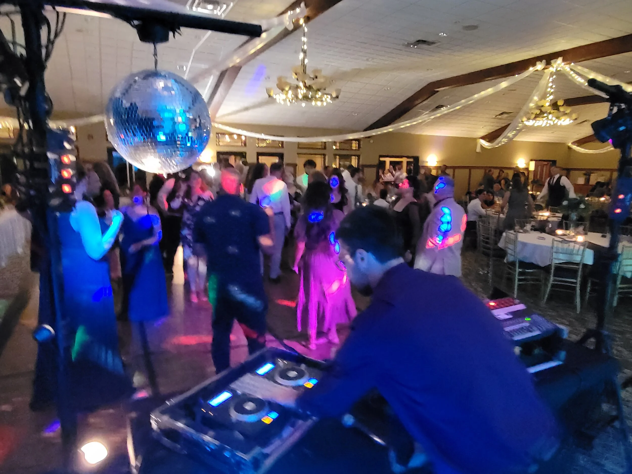 DJ playing music at a lively party with a disco ball, colorful lights, and people dancing on the dance floor in a decorated hall.