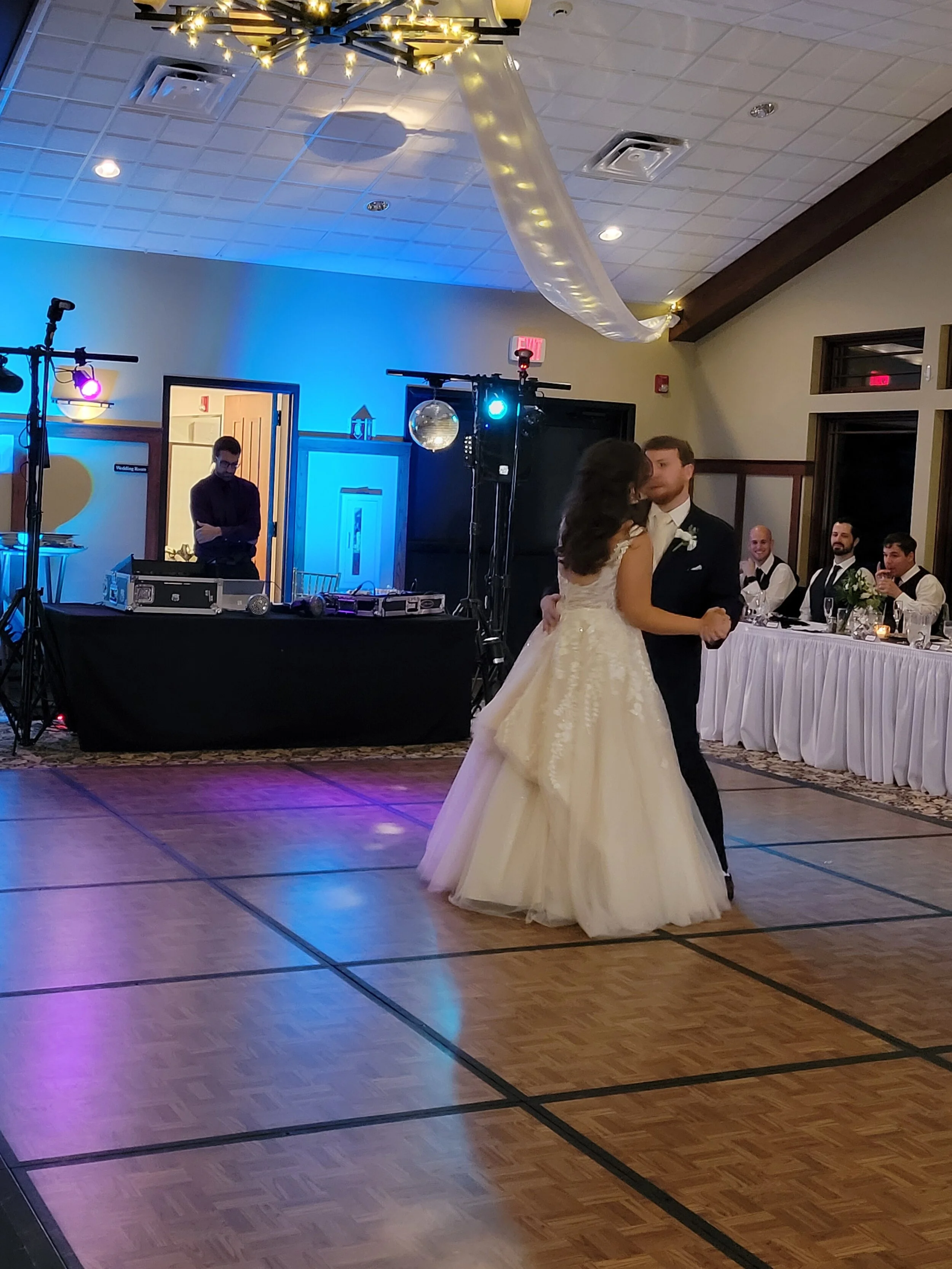 Couple dancing at a wedding reception on a wooden dance floor, with ambient lighting and a DJ setup in the background. Guests are seated at a long table, and decorative lights hang from the ceiling.