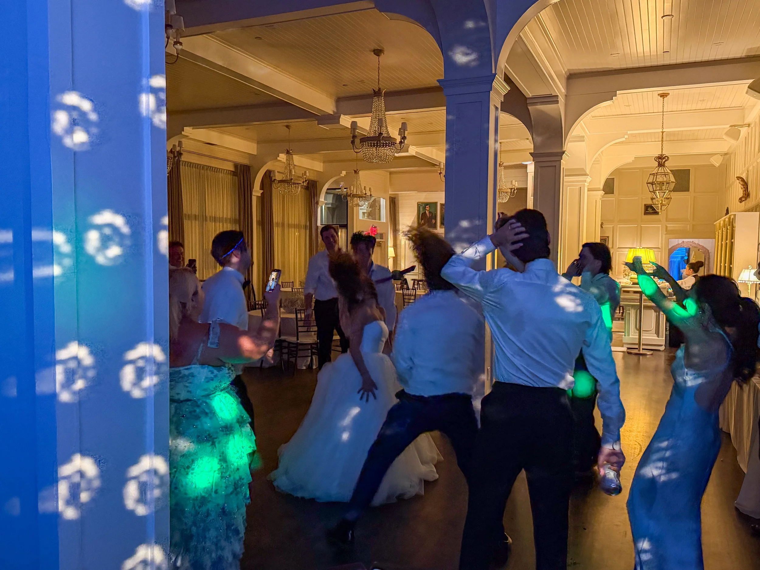 People dancing at a wedding reception inside a decorated banquet hall with chandeliers and portraits on the walls.